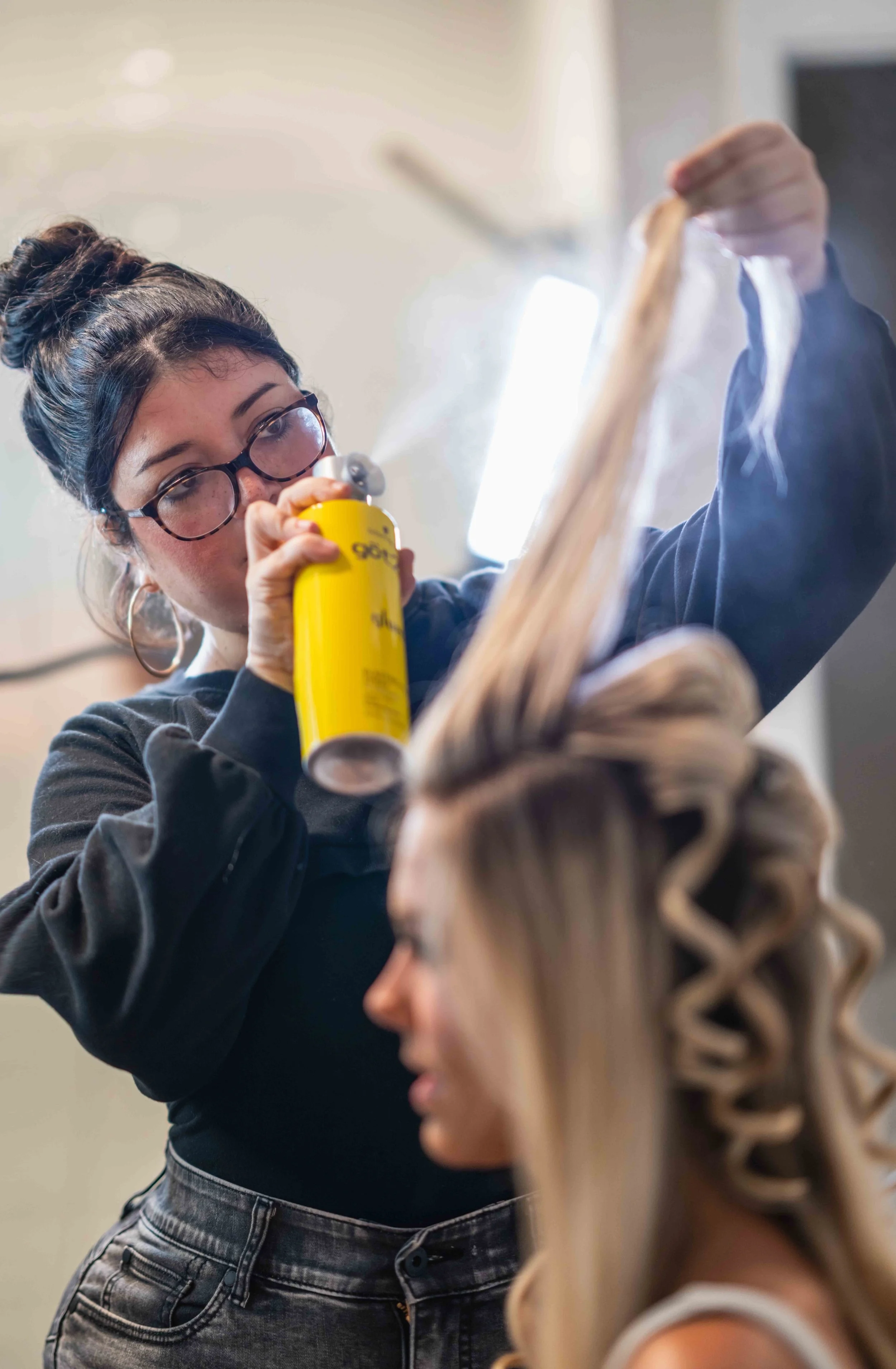A woman wearing glasses sprays hairspray onto a person's hair at a salon or beauty shop. The person getting their hair done has long, curled, blonde hair with a wrap around style. The woman in glasses is focused on her work, holding the hairspray in one hand and a section of hair in the other.