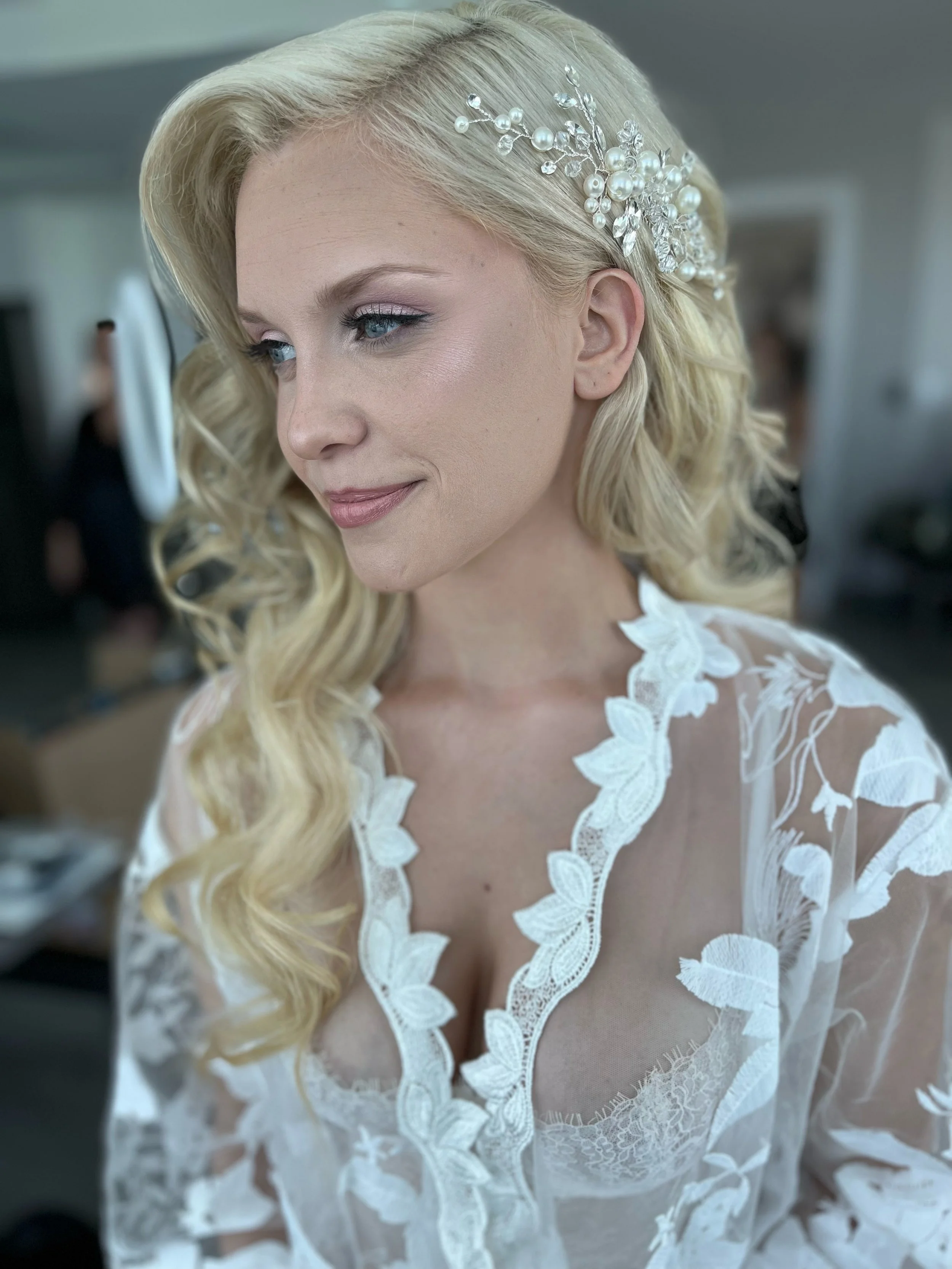 Close-up of a woman with long blonde curly hair, wearing a pearl and crystal hair accessory, and a white lace embroidered dress.
