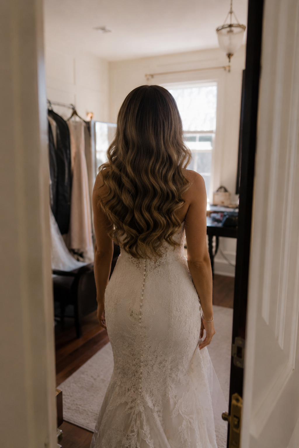Woman in a wedding dress with long wavy hair standing in a room looking out a window.