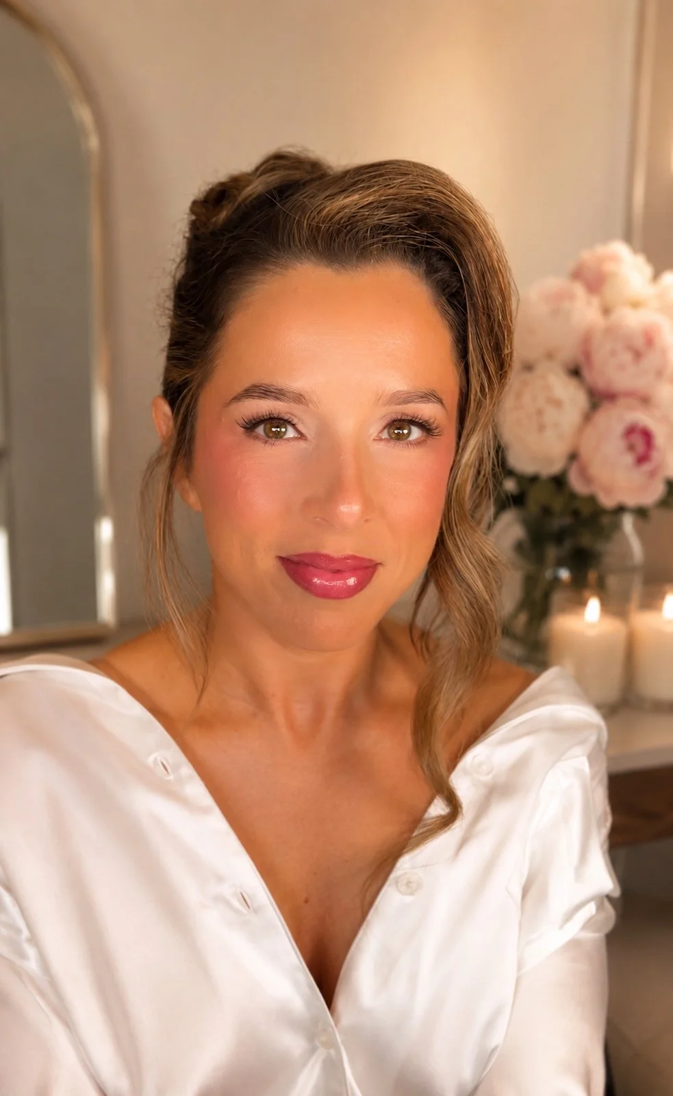 A woman with styled hair, makeup, and a white satin blouse, in a room with a mirror, pink flowers, and lit candles in the background.