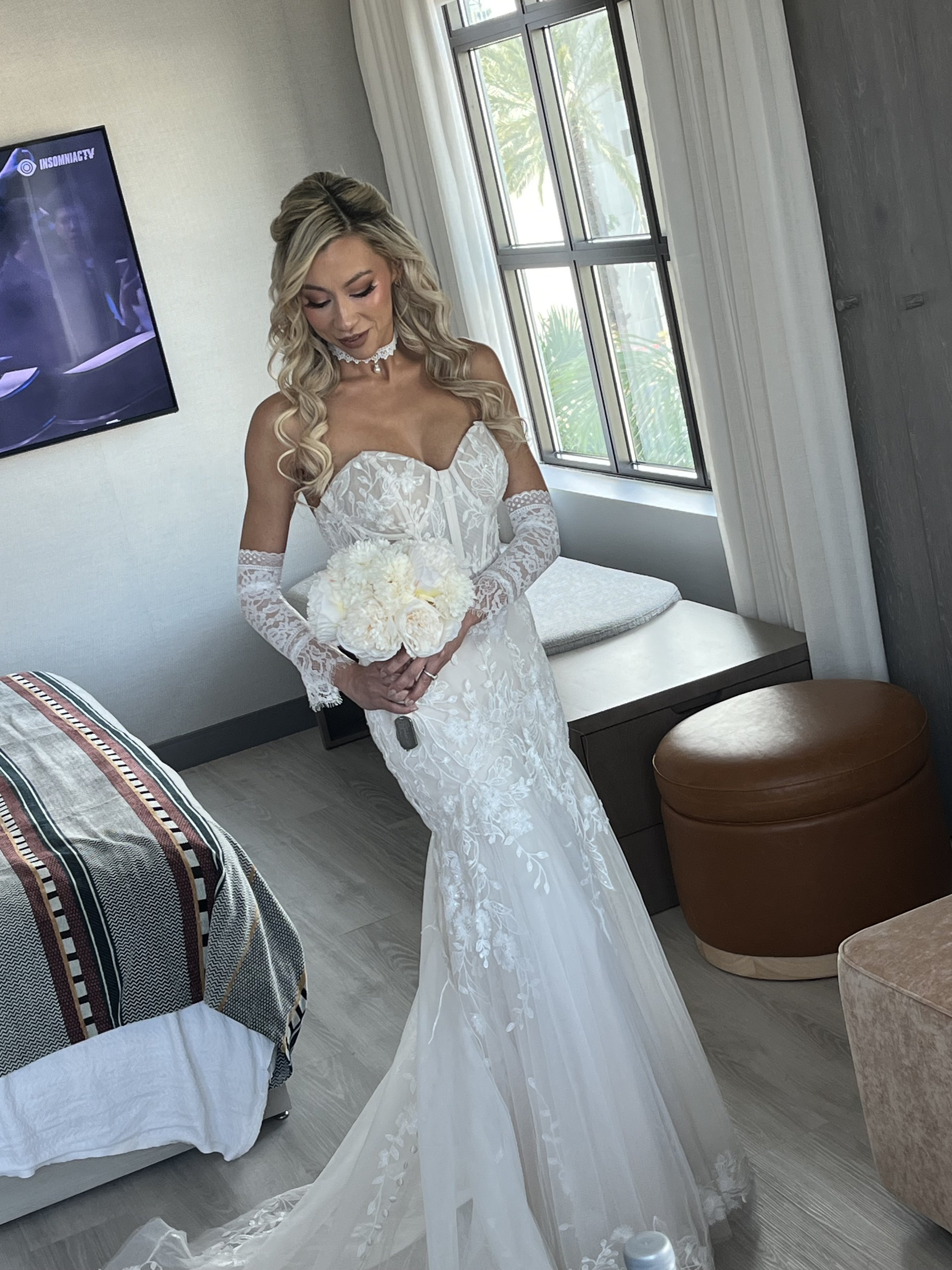 A woman in a white wedding dress with lace and embroidery, holding a bouquet of white flowers, standing in a hotel room near a window with white curtains. She has blonde curly hair and is wearing a choker necklace.