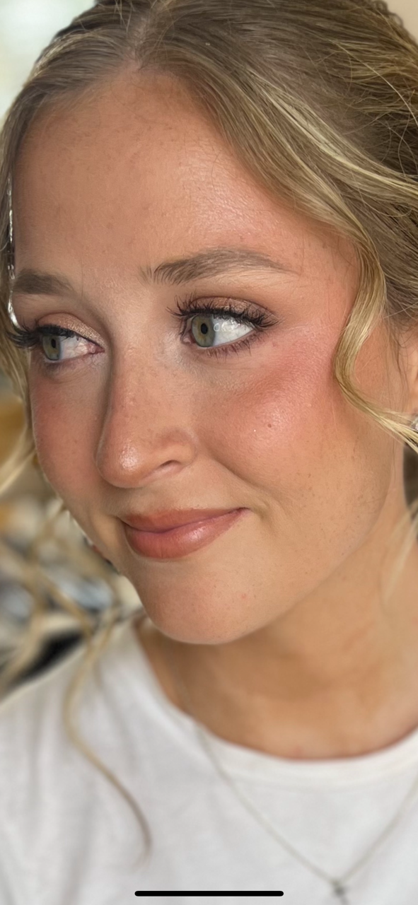 Close-up of a woman with blonde hair styled in soft waves, wearing makeup with natural tones, and a white shirt.
