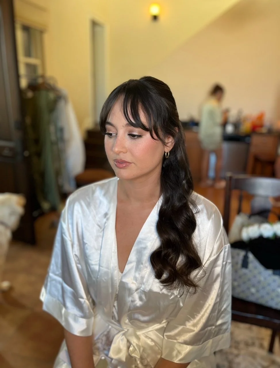 A woman with long dark hair styled in loose waves, wearing a silky white robe, sitting in a room with a blurred background of a person and furniture.