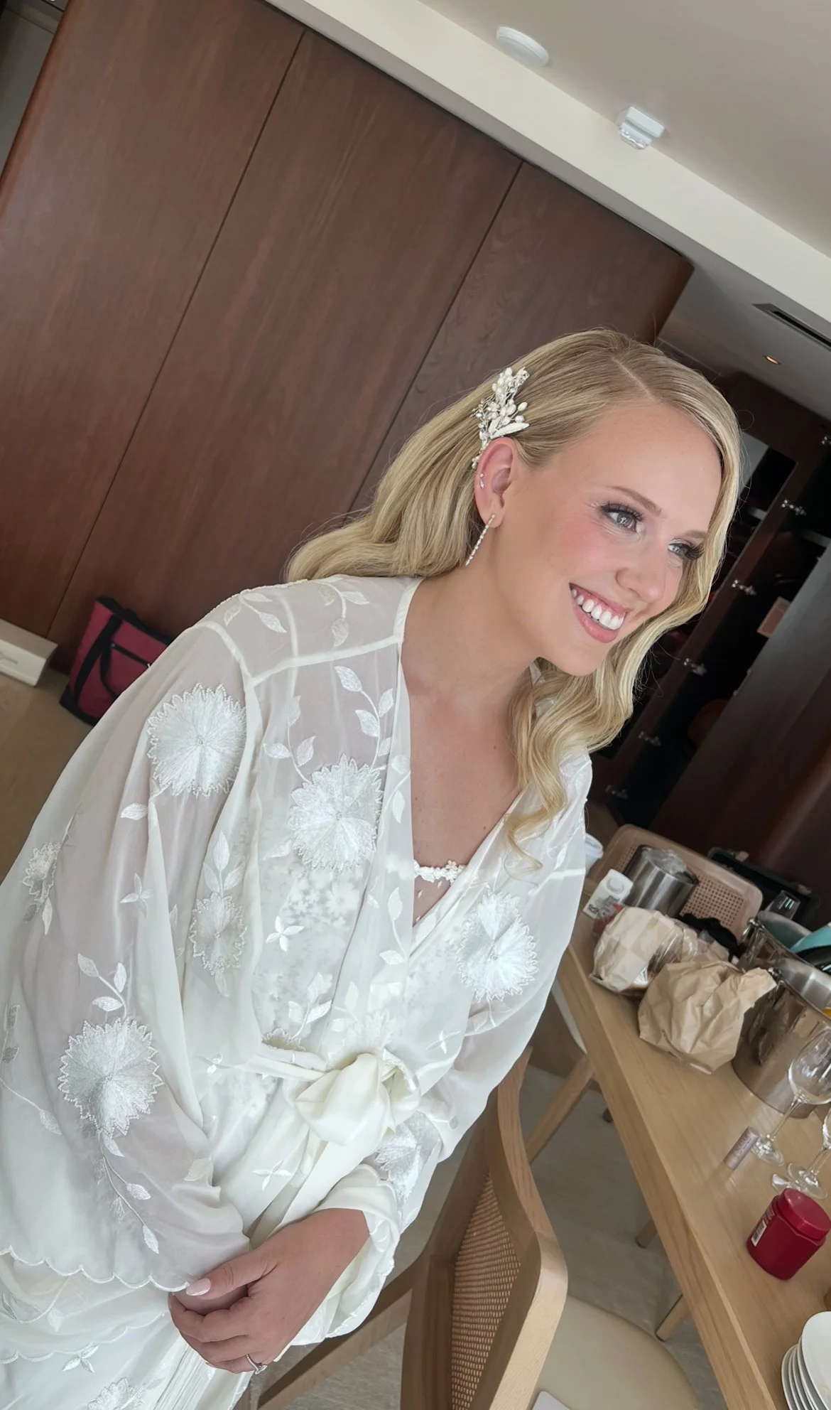 A woman with blonde hair wearing a white embroidered robe and a floral hair clip, smiling and sitting in a room with a wooden cabinet and a table with paper bags, dishes, and cups.