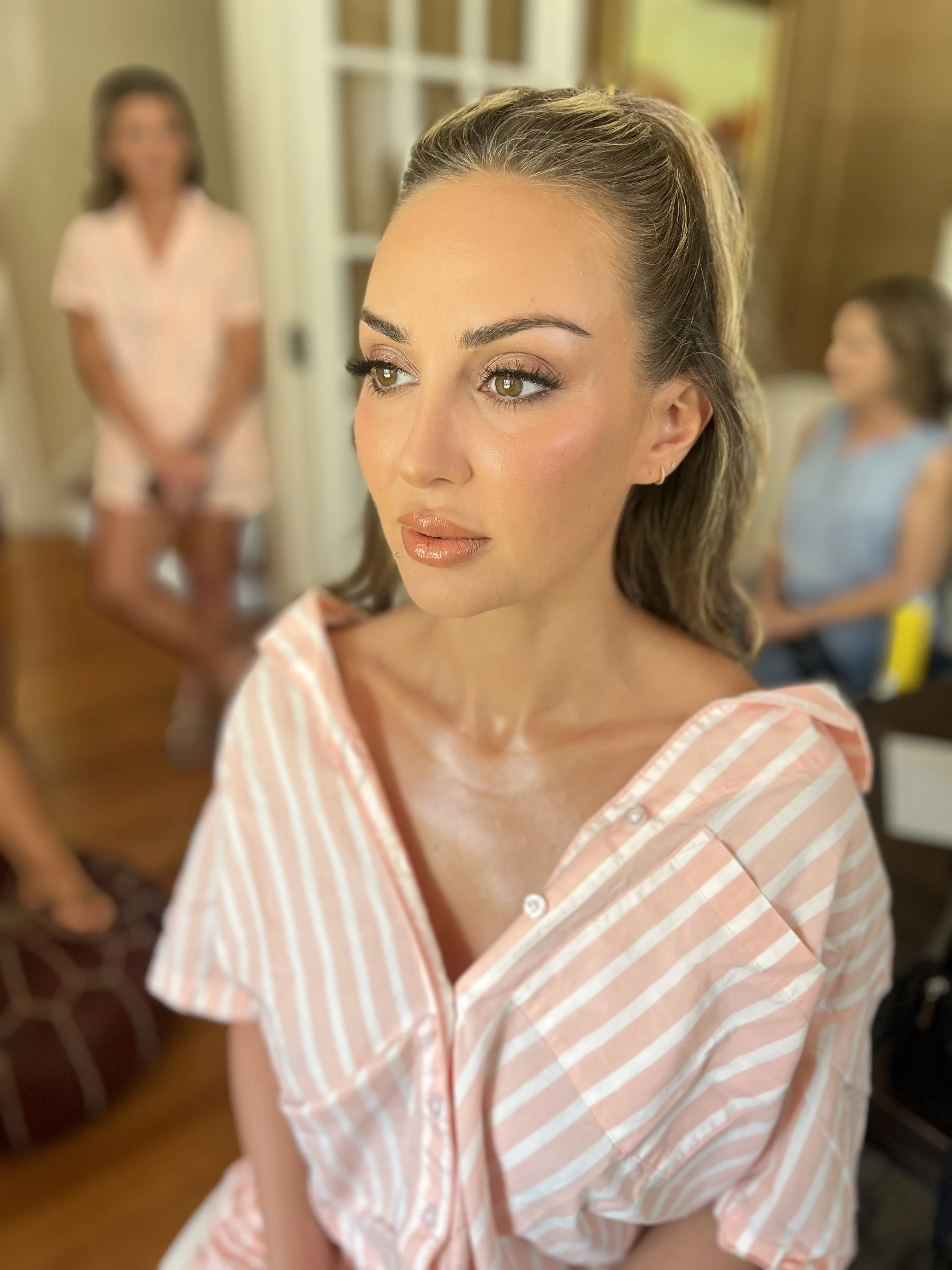 A woman with light face makeup, glossy lips, and brown eyes looking off to the side, wearing a pink and white striped shirt with the top buttons undone. In the background, two other women are visible, one with dark hair in a pink outfit standing with her hands clasped and another with lighter hair in a blue top, seated and smiling.