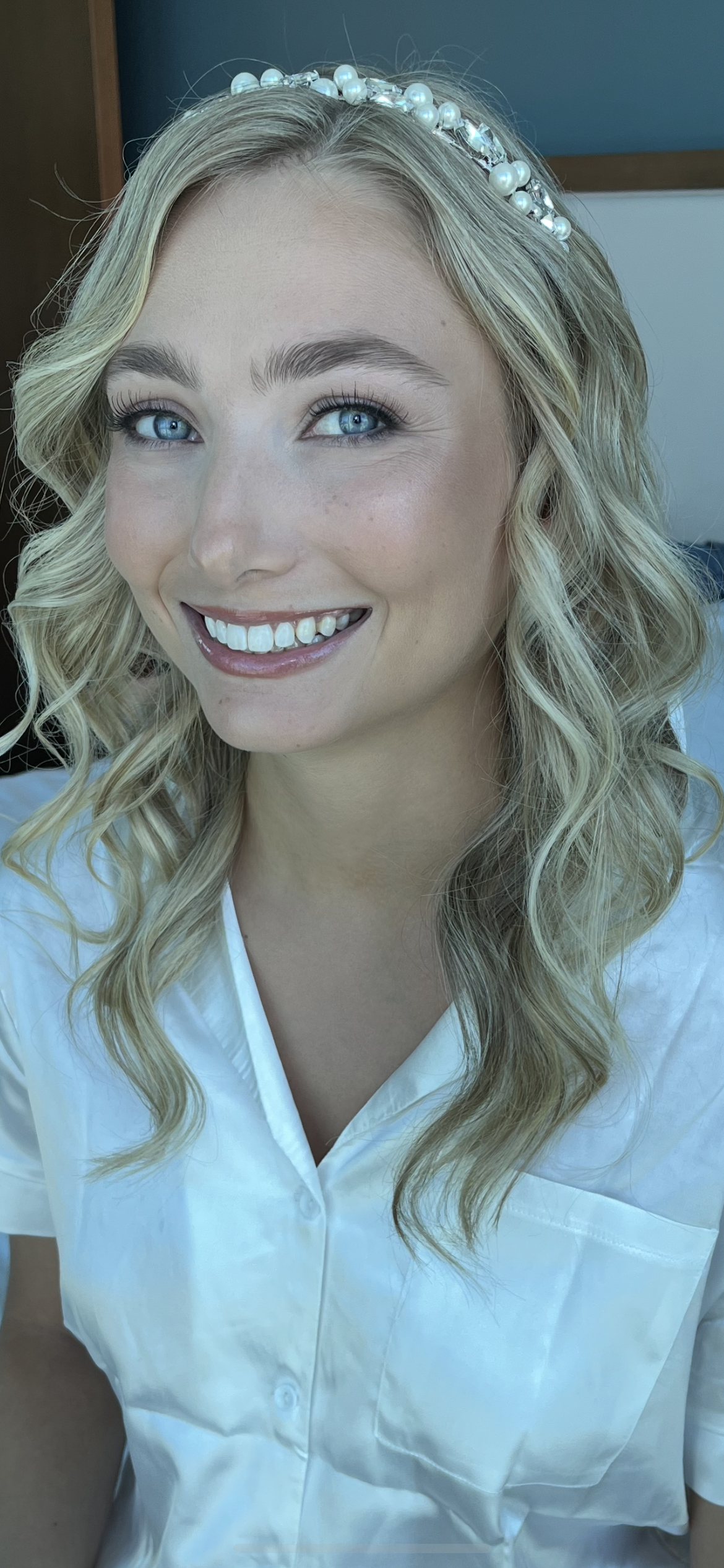 Close-up of a woman with blonde wavy hair, blue eyes, wearing a pearl headband and a white shirt, smiling.