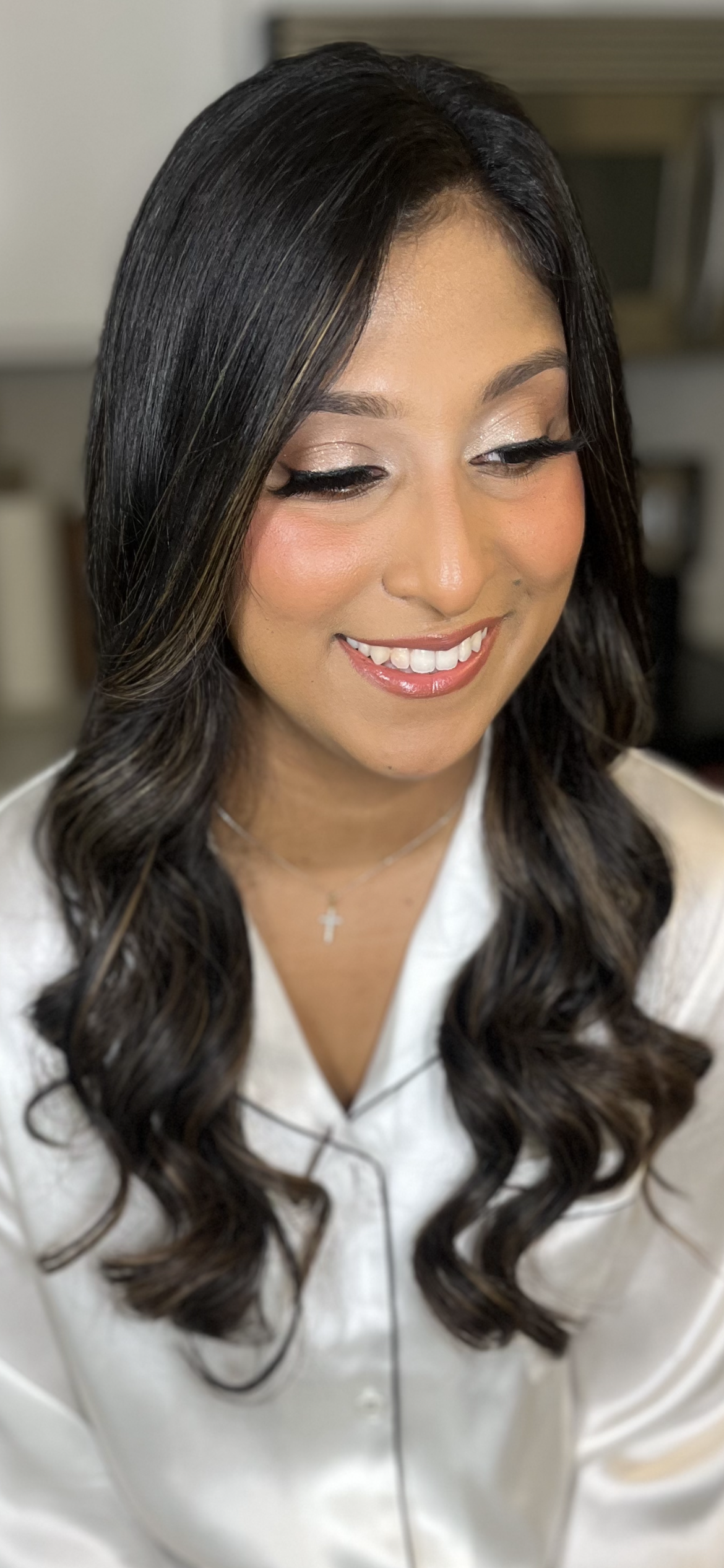 A woman with long dark hair styled in loose curls, wearing makeup with shimmery eyeshadow and glossy lips, smiling softly, dressed in a white top with a cross necklace.