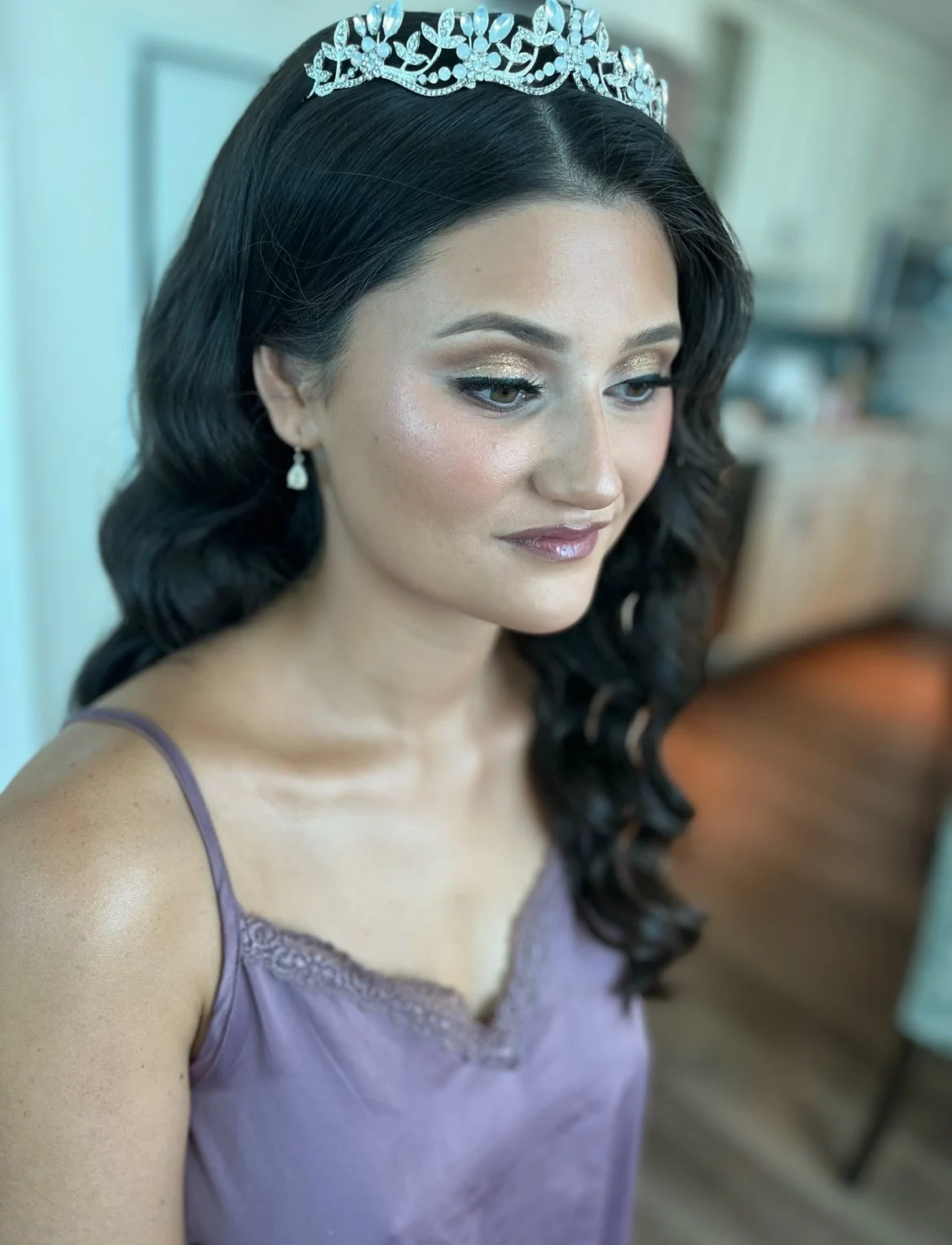 Women with dark curly hair wearing a lavender dress with lace details, a silver tiara, and teardrop earrings, looking to the side.