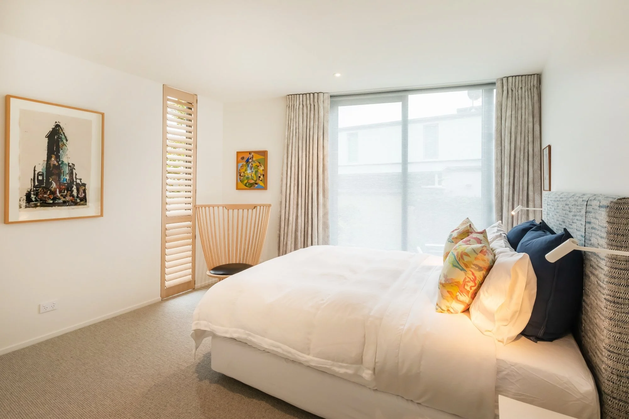 A bedroom with a white bed, colorful pillows, a wooden chair, art on the walls, large windows with curtains, and outdoor view.