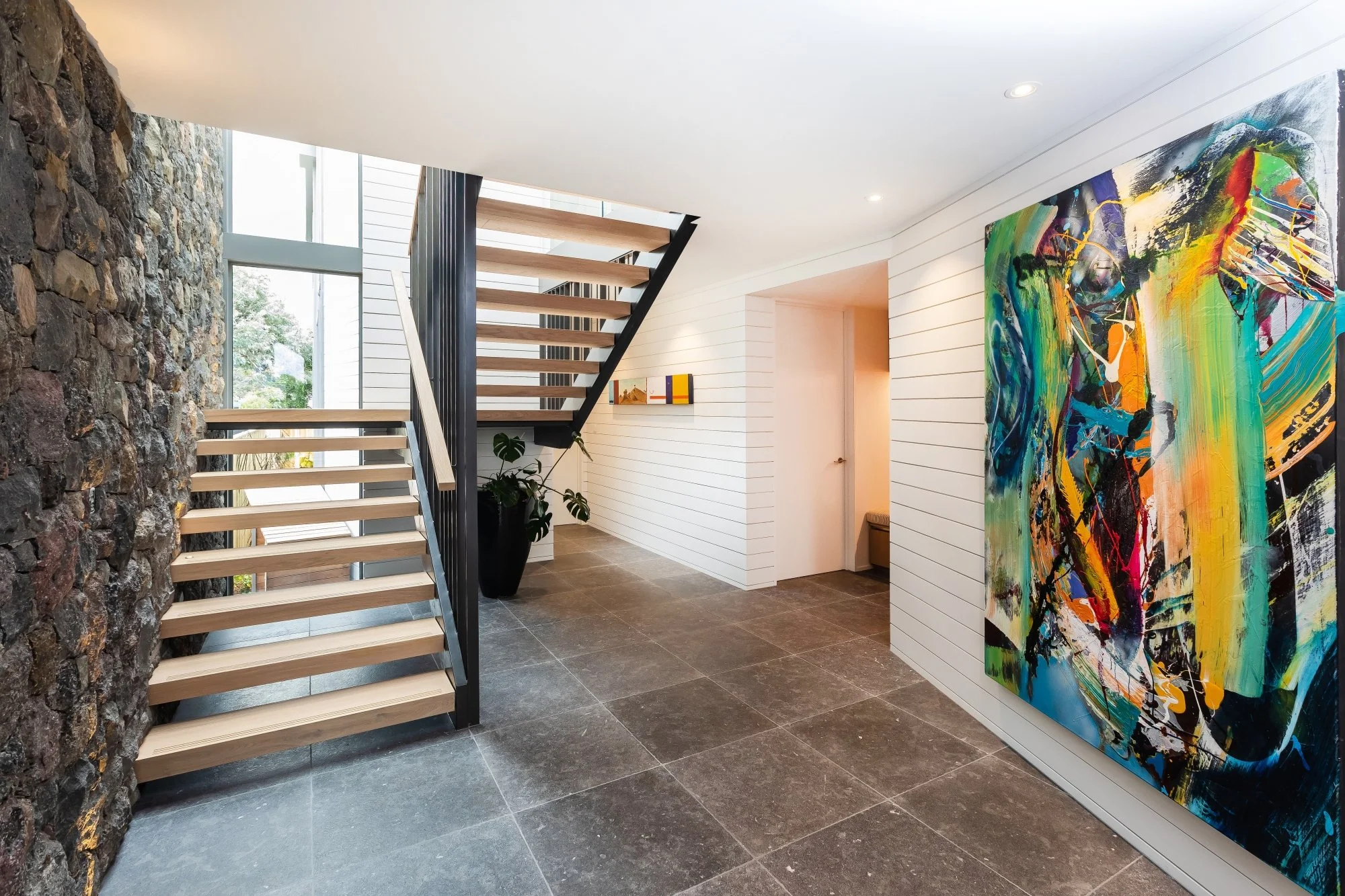 Modern interior stairway with wooden steps and black railings, stone accent wall on the left, tall windows letting in natural light, colored abstract art on white shiplap wall, and potted plant near the wall. Chamoille Gris stone to the floor.