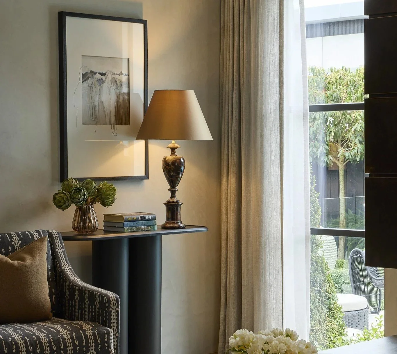 A cozy living room corner with a patterned armchair, a side table holding a vase with green flowers, bounded books, a decorative table lamp, a framed line art portrait on the wall, designer fabric double layer curtains, 