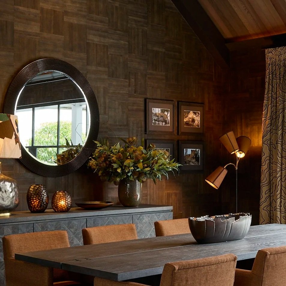 A dining room with a dark wooden wall and matching ceiling. A large oval mirror is on the wall, reflecting a window with greenery outside. Below the mirror is a sideboard holding a large floral arrangement and decorative vases and bowls. A framed set