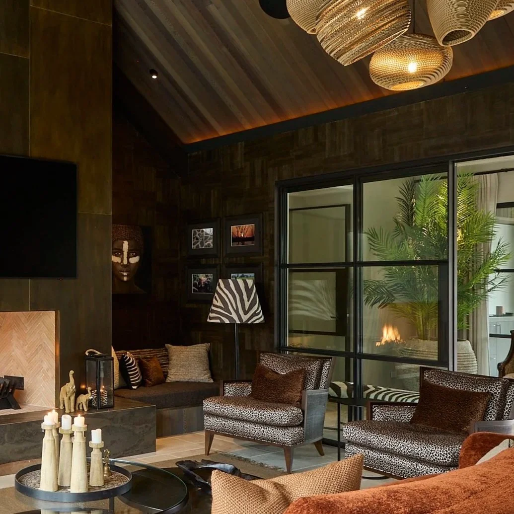Living room with dark wood walls, a fireplace, patterned chairs, framed photos, a large window, and hanging light fixtures.