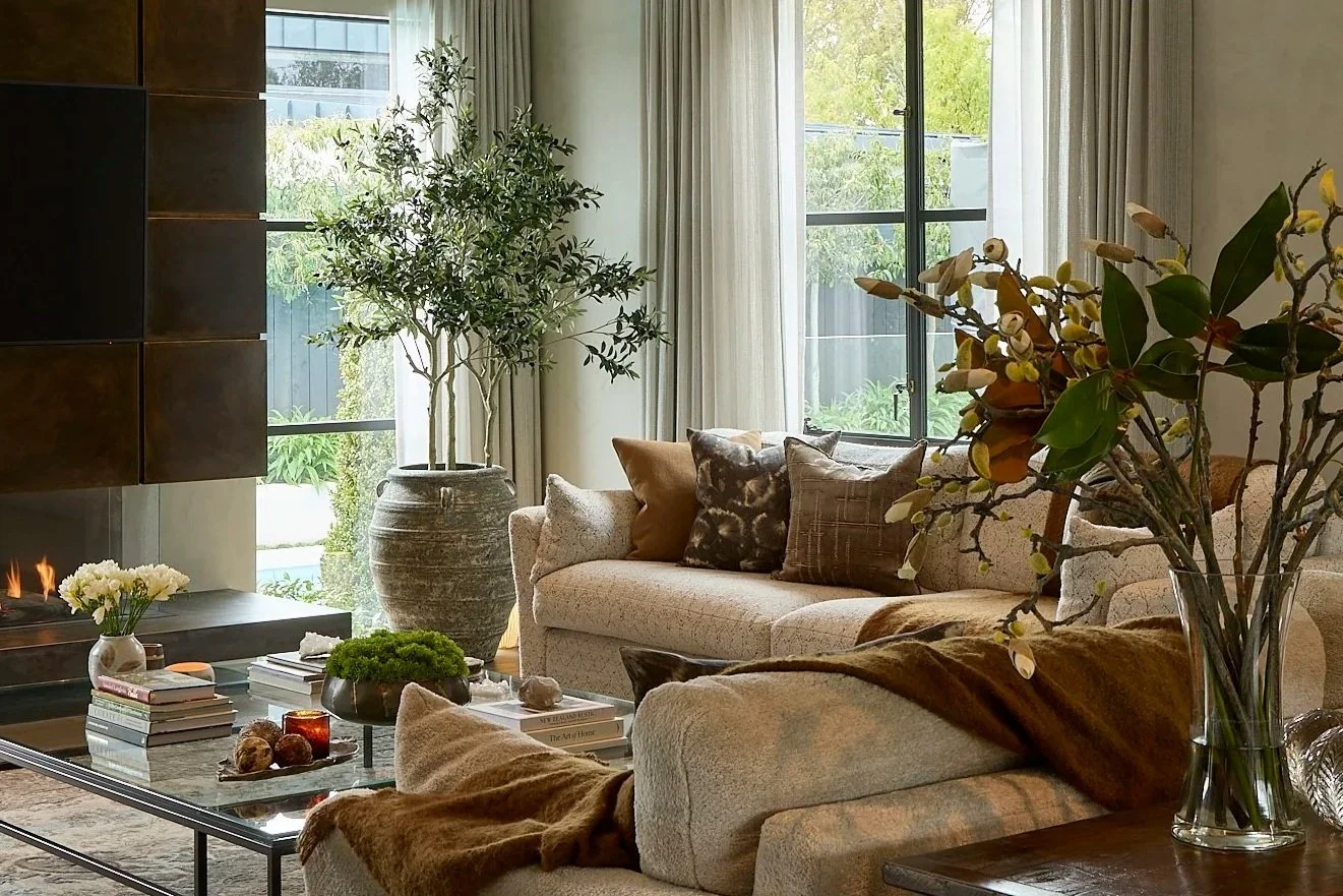 A cozy living room with a beige sofa decorated with various patterned and solid throw pillows, a glass coffee table with books, candles, and decorative items, a large potted plant near the window, and a vase with flowers on a side table. Large window