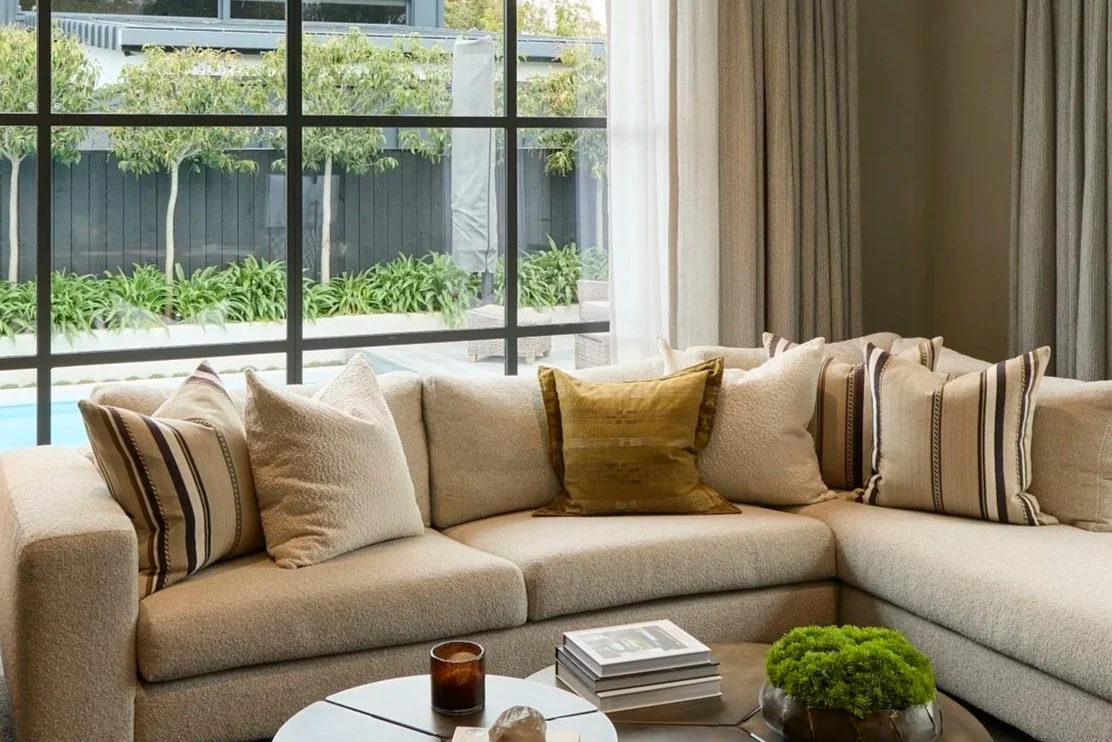 Living room with a beige boucle sectional sofa, with striped Larsen cushions. a round brass coffee table with magazines, a small green plant, and a brown candle. Large windows with curtains reveal a backyard garden with green plants and trees.