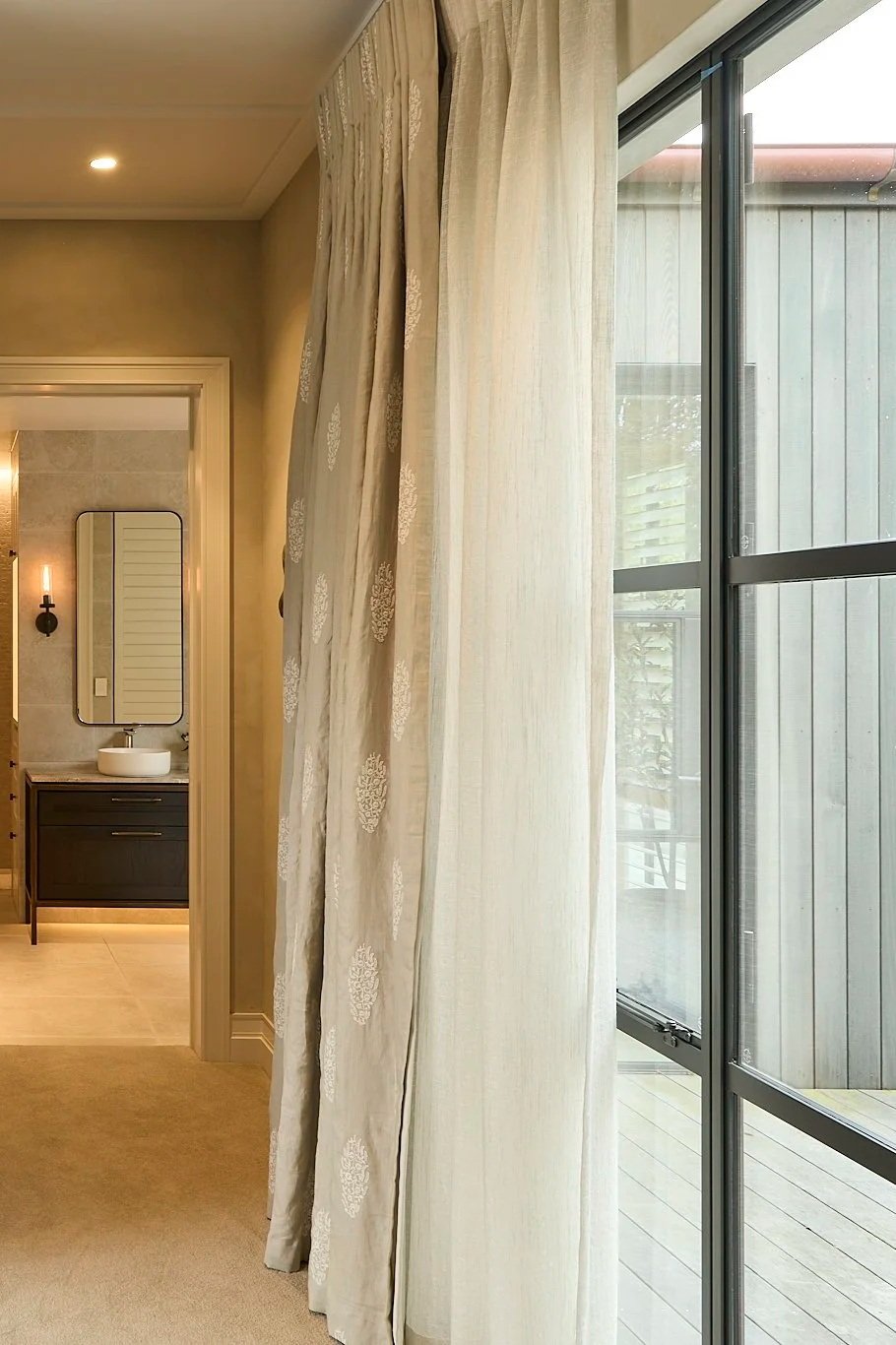 View of a living room with soft sheer curtains on doule bronze rod and a sliding glass door leading to a wooden deck outside, with a bathroom vanity with a mirror and vessel sink in the background.