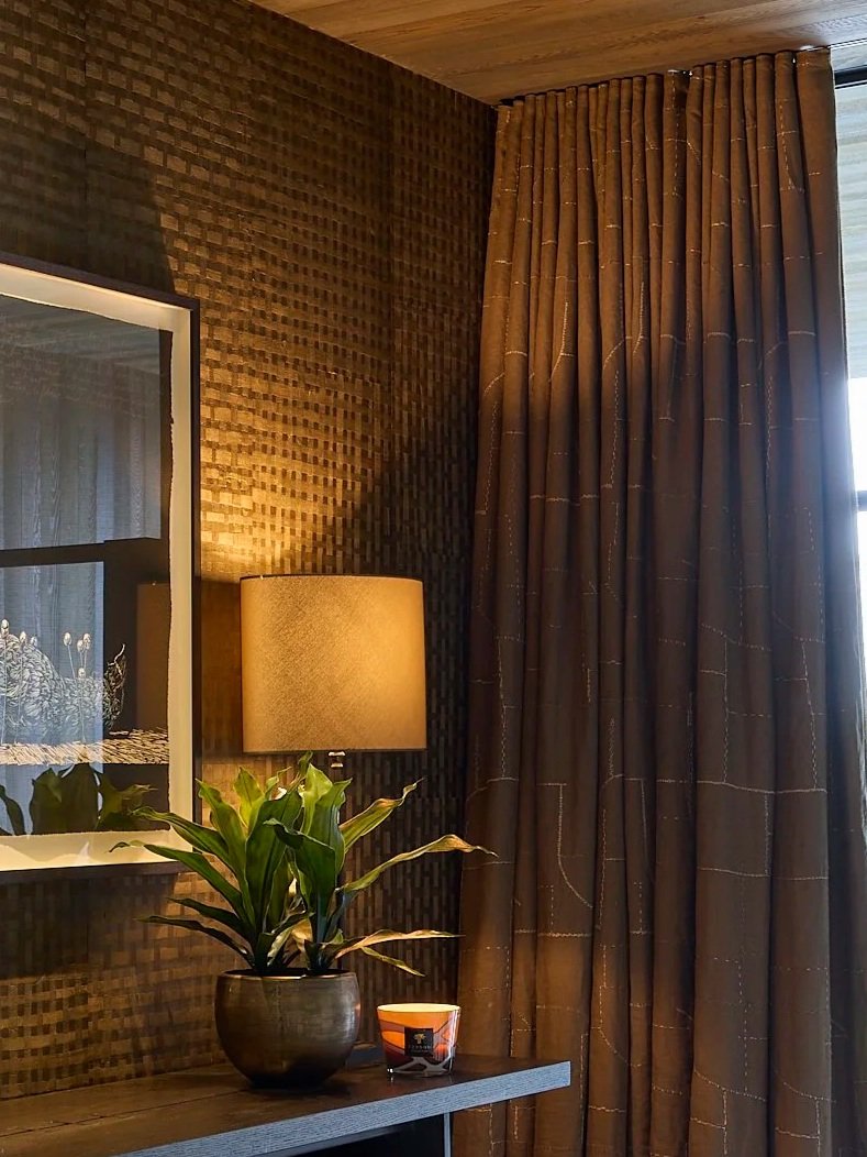 Inside a room with a textured brown wall, a beige lampshade on a lamp, a green plant in a dark pot on a table, a framed picture on the wall, and designer embroidered wave top curtains covering a window.