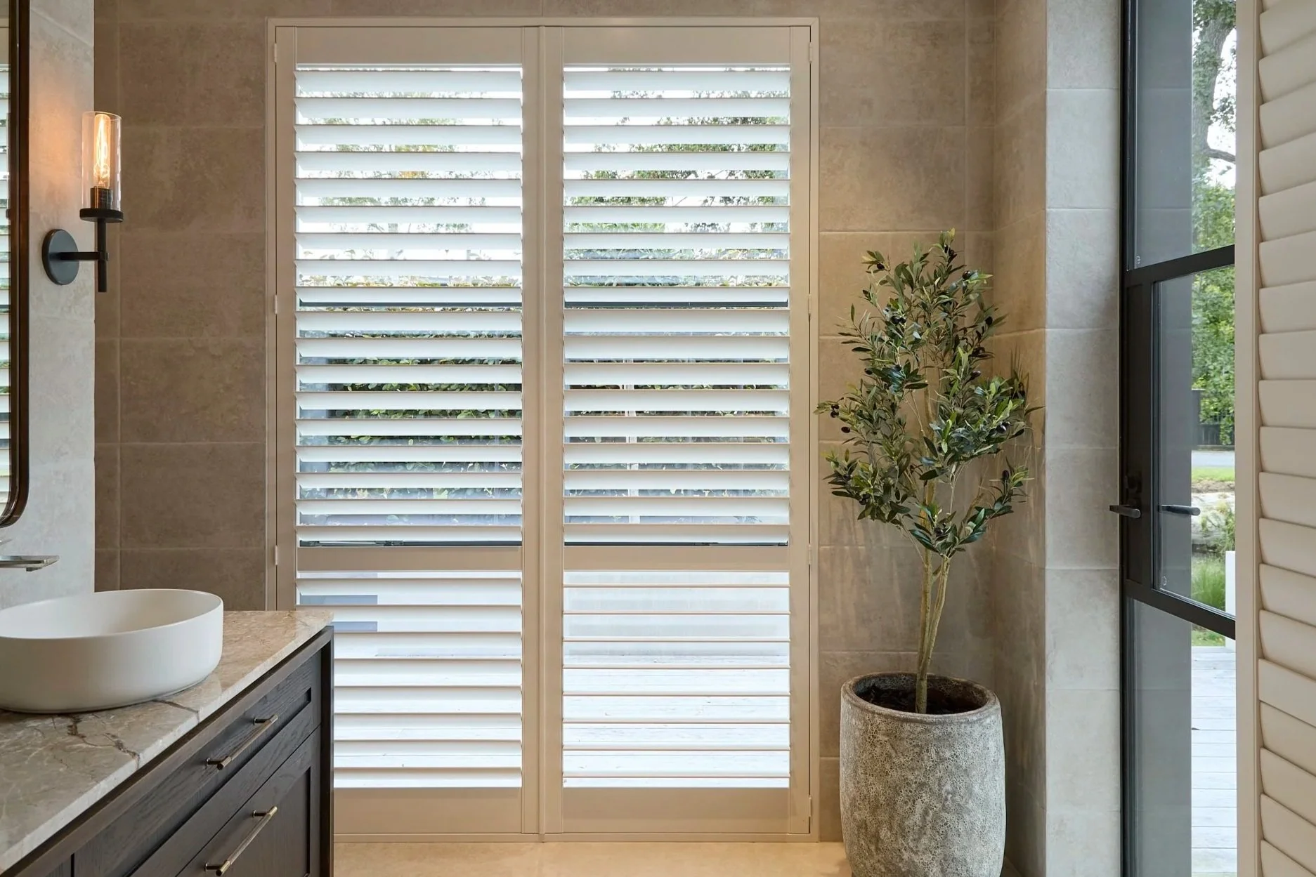 Modern bathroom with beige tiled walls, a white louvered window, a black vanity with a white vessel sink, a wall-mounted light fixture, a potted olive tree, and a glass door leading outside.Norman white shutters sliding and hinged.