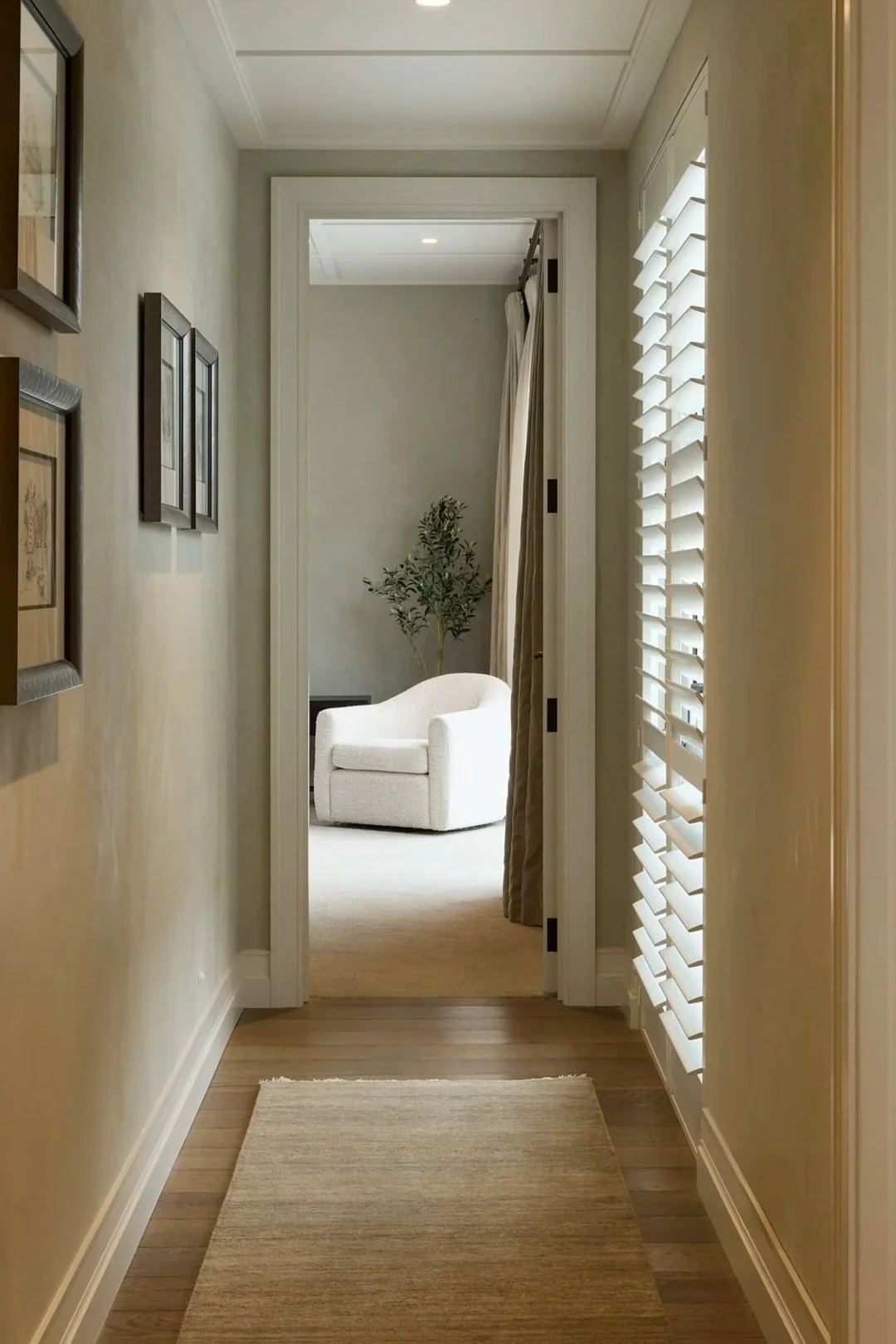 Armadillo Odessa rug runner on floor. and Norman white Shutters  to the window. A hallway leading to a room with a white armchair and a potted plant, with window shutters on the right and framed pictures on the left wall.