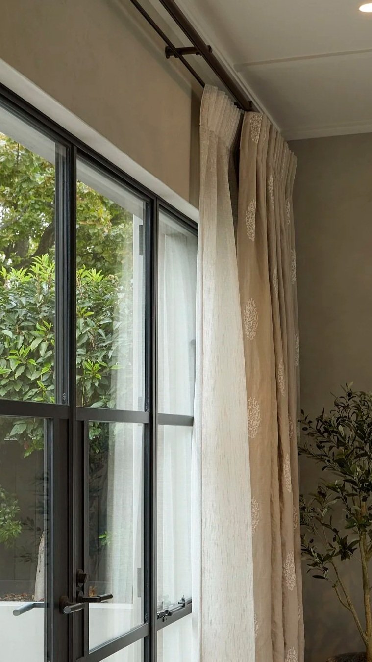 Close-up of a sliding glass door with double tracked sheer linen curtains and a view of green foliage outside.Seneca