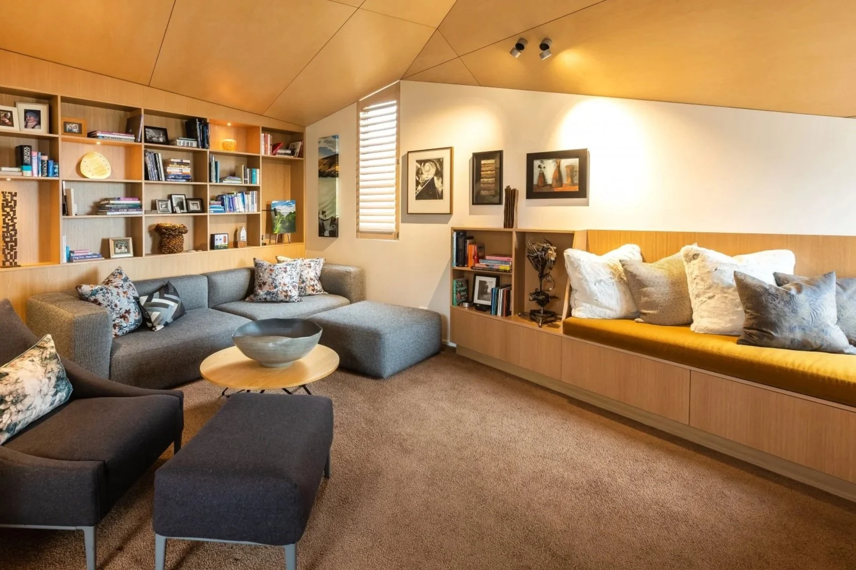 A cozy living room with a gray sectional sofa and matching ottoman, a black armchair with floral pillows, a round wooden coffee table with a decorative bowl, a built-in wooden bookshelf on the wall filled with books and decor, a window with white bli