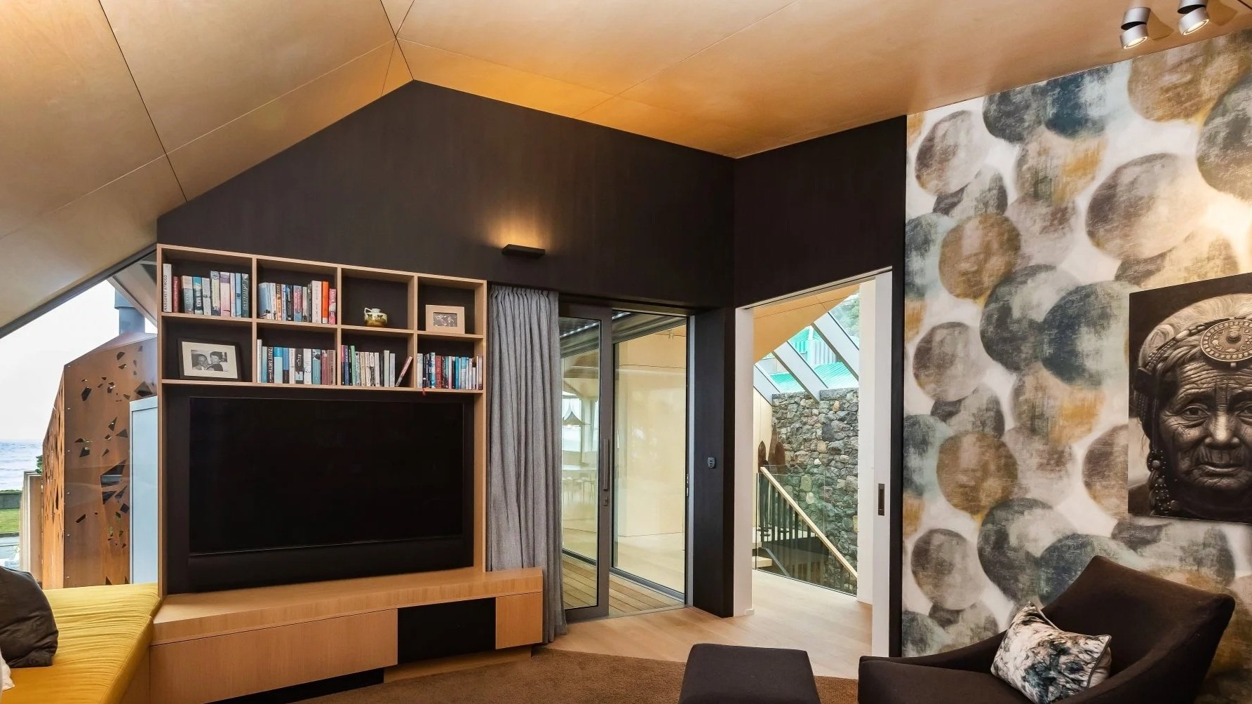 Living room with a large flat-screen TV, a bookshelf filled with books and framed photos, a sliding glass door with gray curtains, a wall with stone and abstract patterned wallpaper, and a sculpture of an elderly woman's face with traditional headgea