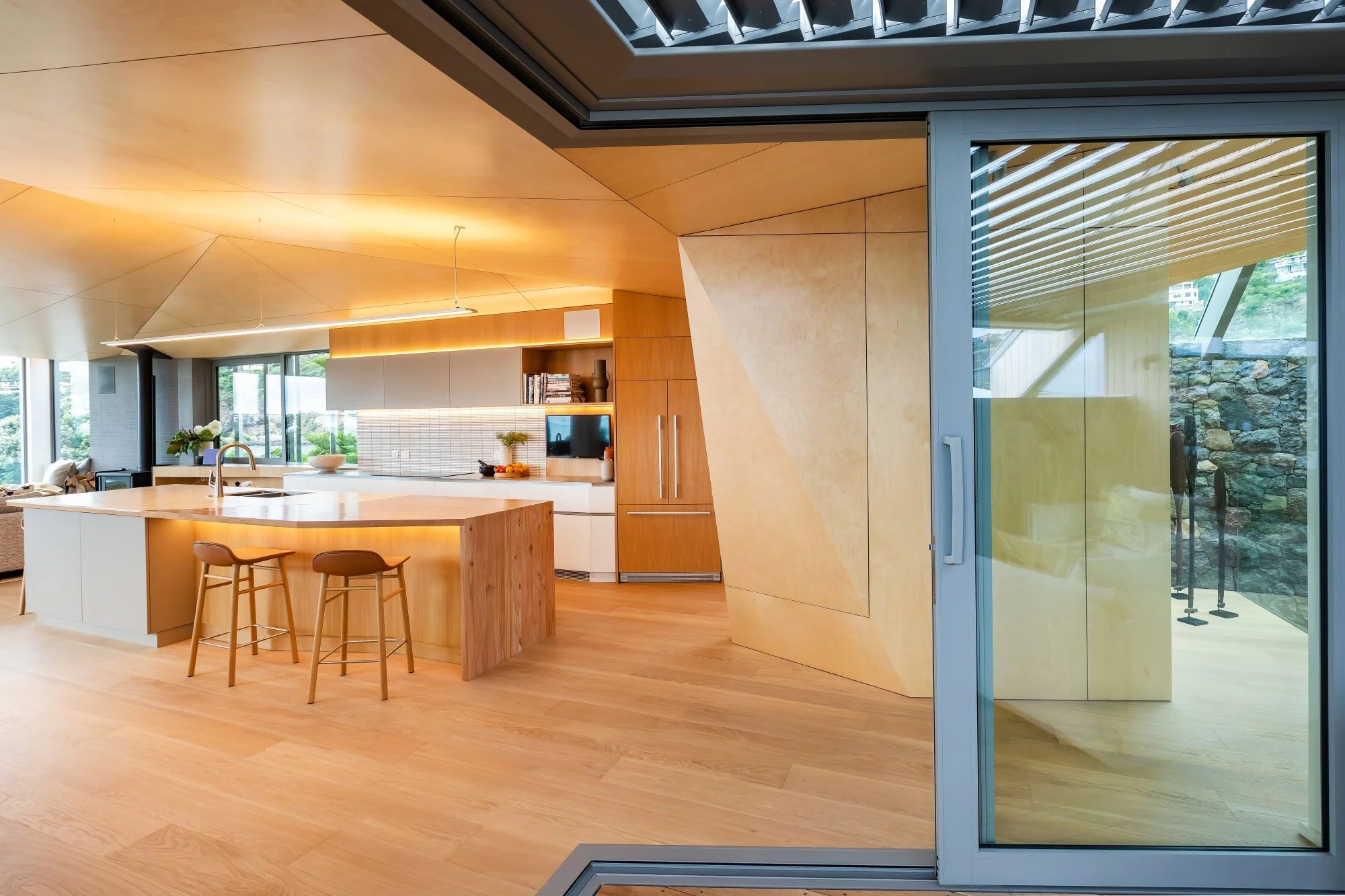 Modern open-concept kitchen and living area with wooden accents, large windows, and a sliding glass door leading to an outdoor space with rocks and greeneryInterior design Bridget Coles Interior Design Christchurch BCSTUDIO