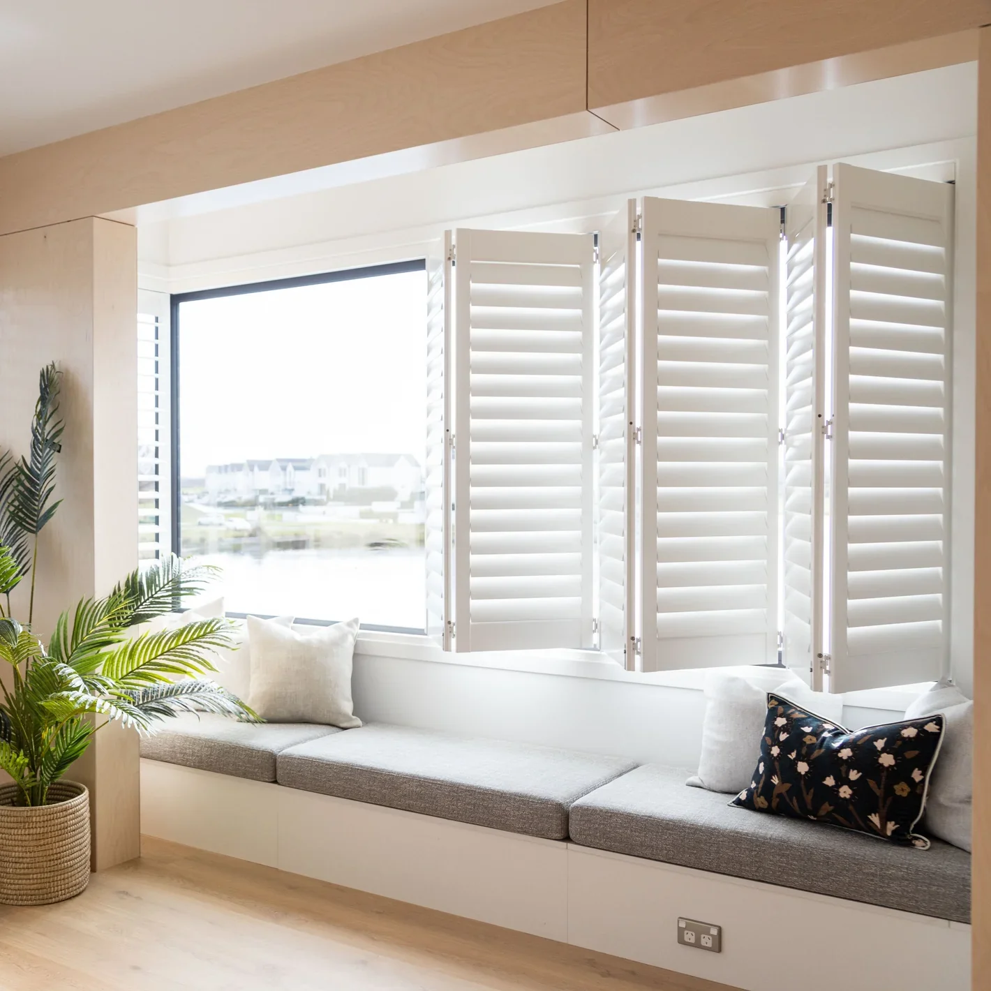 Bright room with large window and Norman white plantation bifold shutters, built-in window seat with upholstered window seat gray cushion and pillows, potted plant,