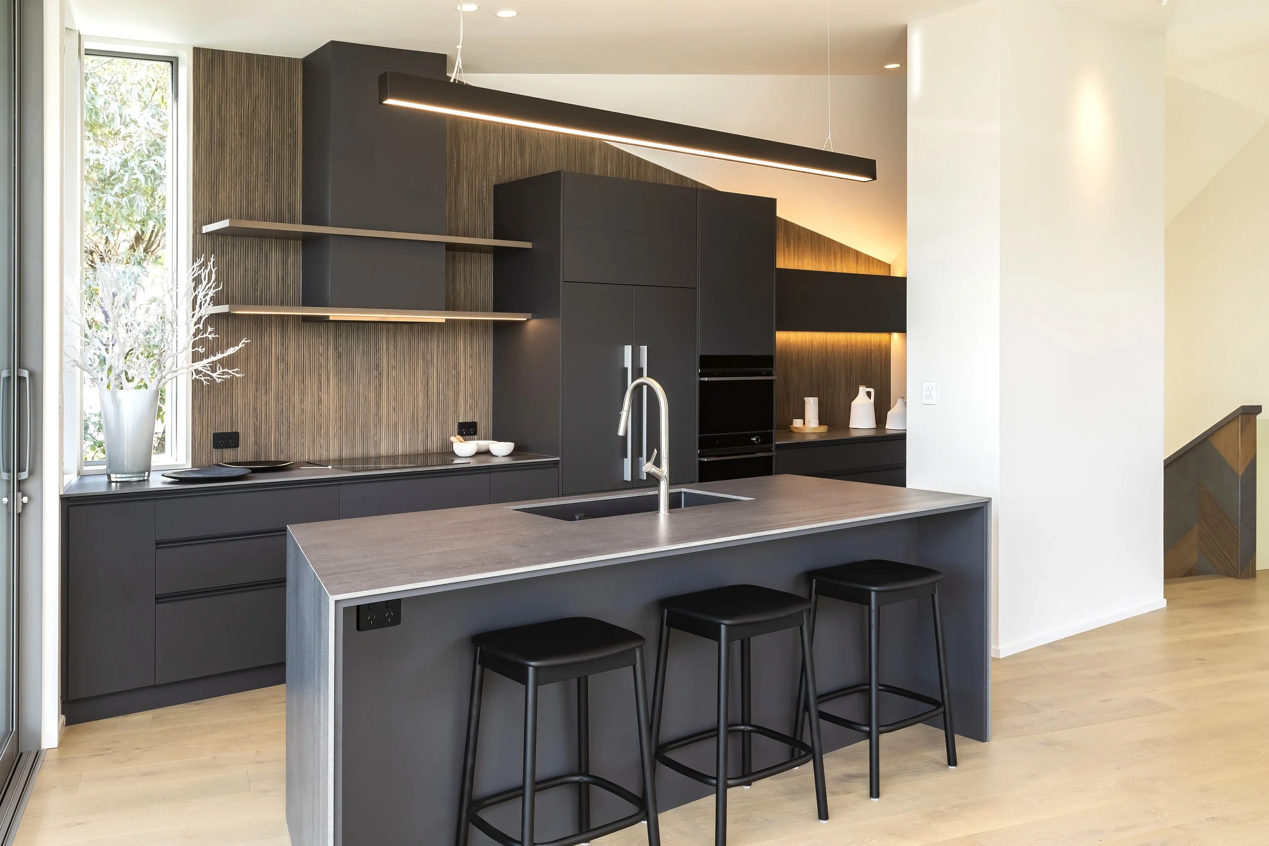 Modern kitchen with black cabinetry, wooden wall accents, a large kitchen island with a gray countertop and black barstools, open shelves, and decorative vases, with a window letting in natural light. Kitchen Design by Bridget Coles Interior Design