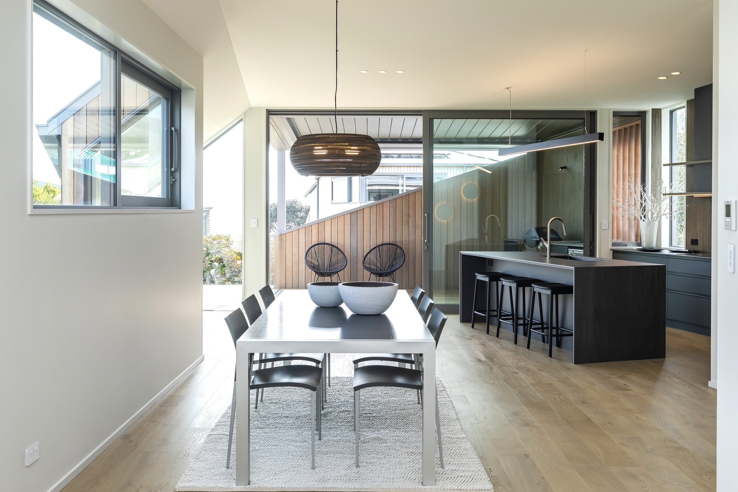 Modern kitchen and dining area with open layout, featuring a white dining table, black chairs, a kitchen island with black barstools, wooden flooring, large windows, and contemporary pendant and linear lighting fixtures. Kitchen Design by Bridget Col