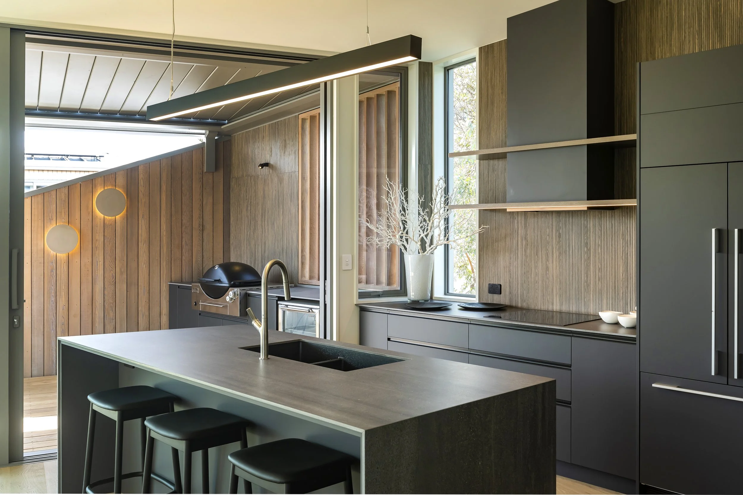 Modern kitchen with a large central island, black bar stools, wooden and gray cabinetry, open shelves, a window with daylight, and decorative elements like a white coral sculpture.