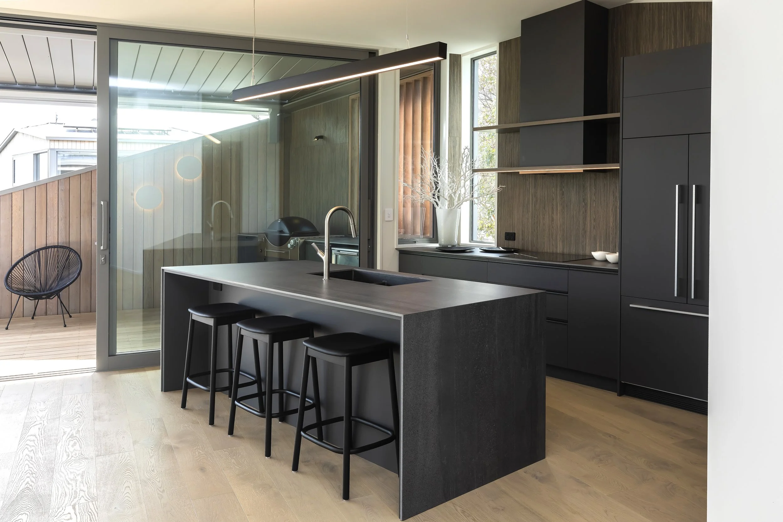 Modern kitchen with black cabinets, a kitchen island with a sink and three black stools, large windows, and a sliding glass door leading to a balcony with wooden flooring and outdoor seating. Kitchen Design by Bridget Coles Interior Design