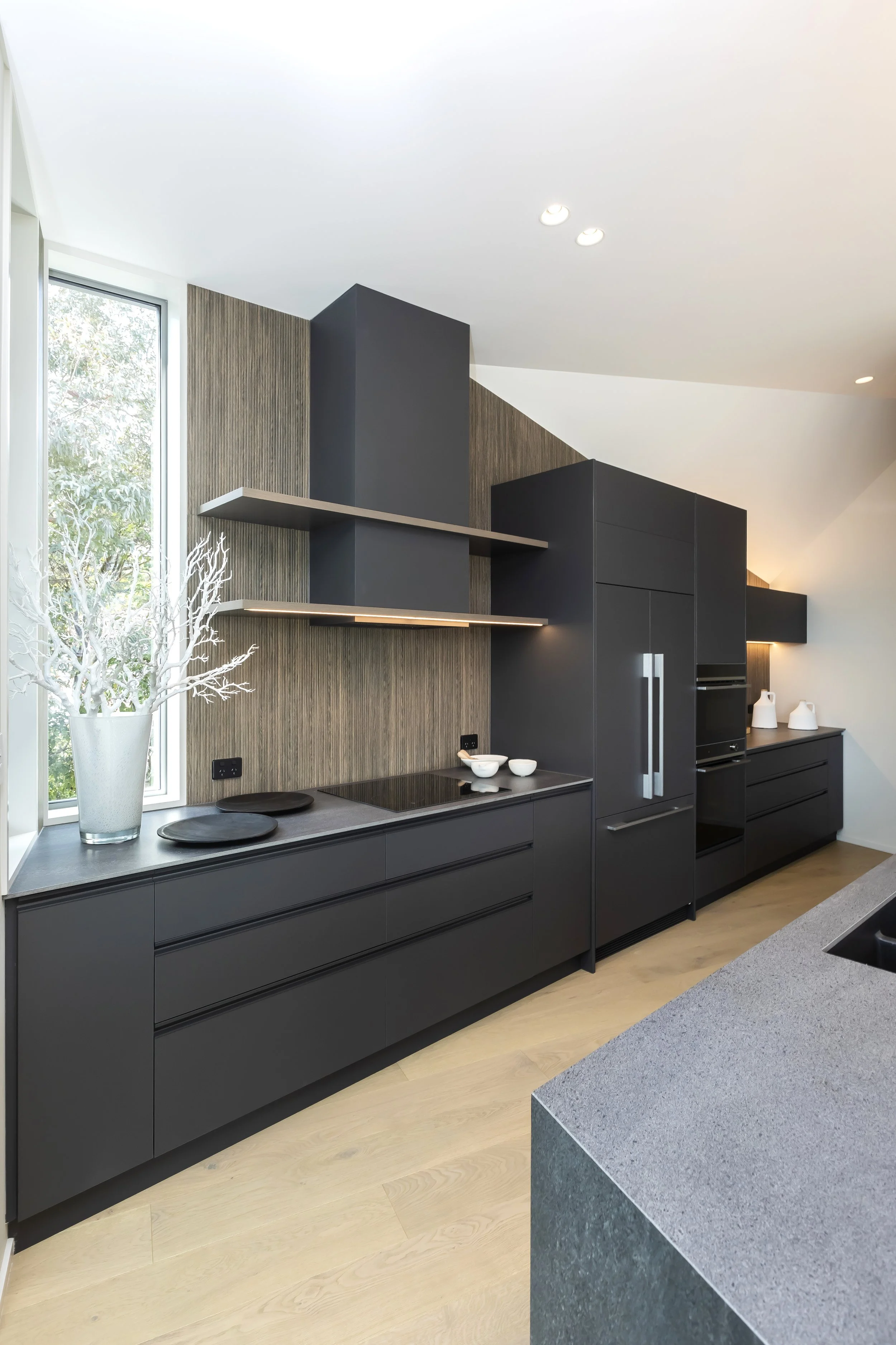 Modern kitchen with black cabinetry, a gray countertop, a large window, and decorative white branches in a vase. Kitchen Design by Bridget Coles Interior Design