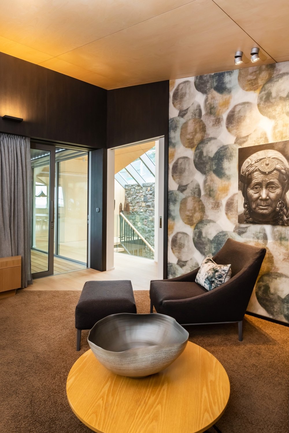 Living room with a black armchair and matching ottoman, a wooden coffee table with a ceramic bowl, large window with curtains, artwork of rocks and an African mask on the wall, and a view of a staircase and stone wall outside.