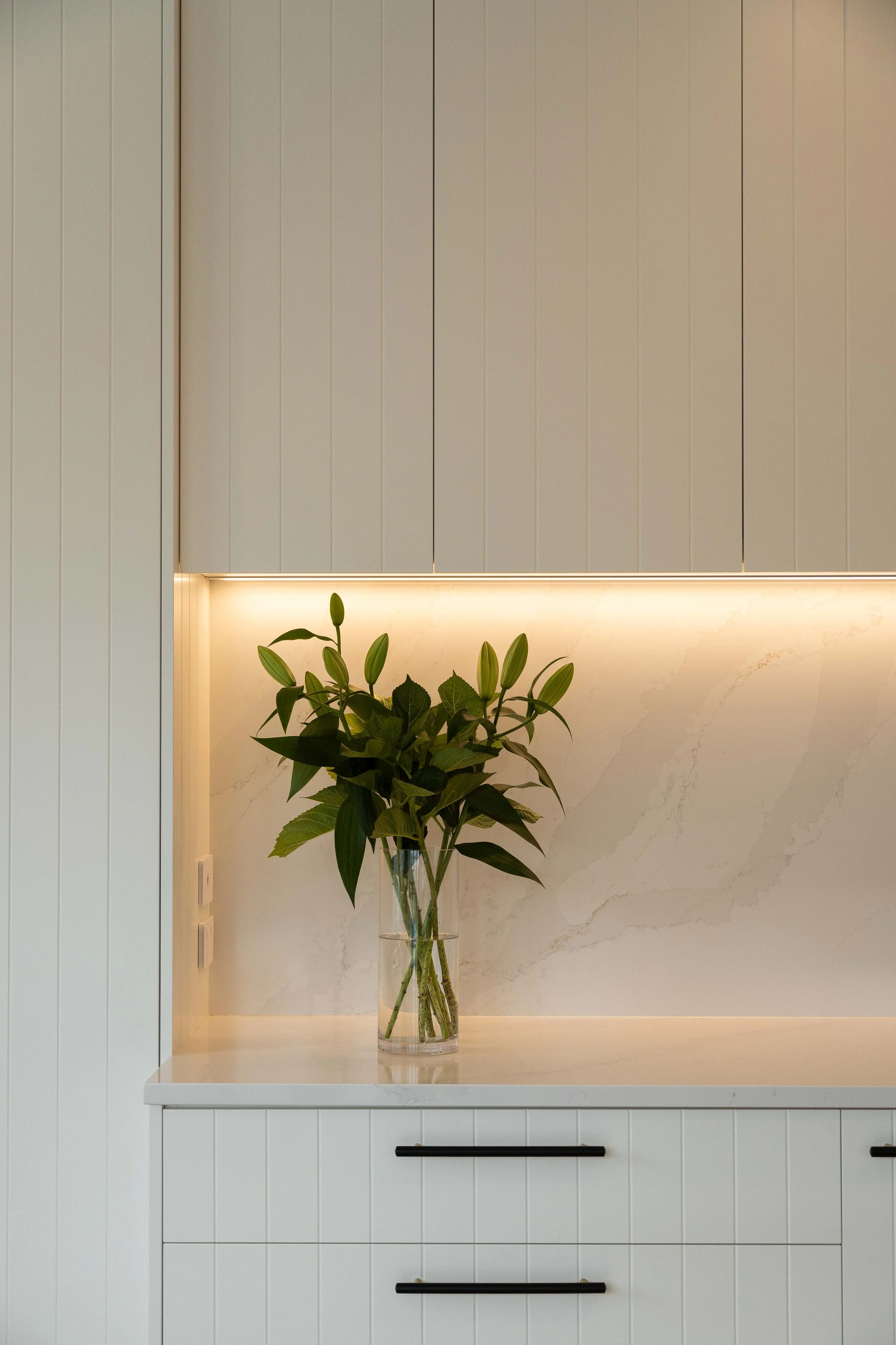 A kitchen or modern interior with white cabinets, a white marble countertop, and a large glass vase holding green leafy plants, illuminated by warm lighting.