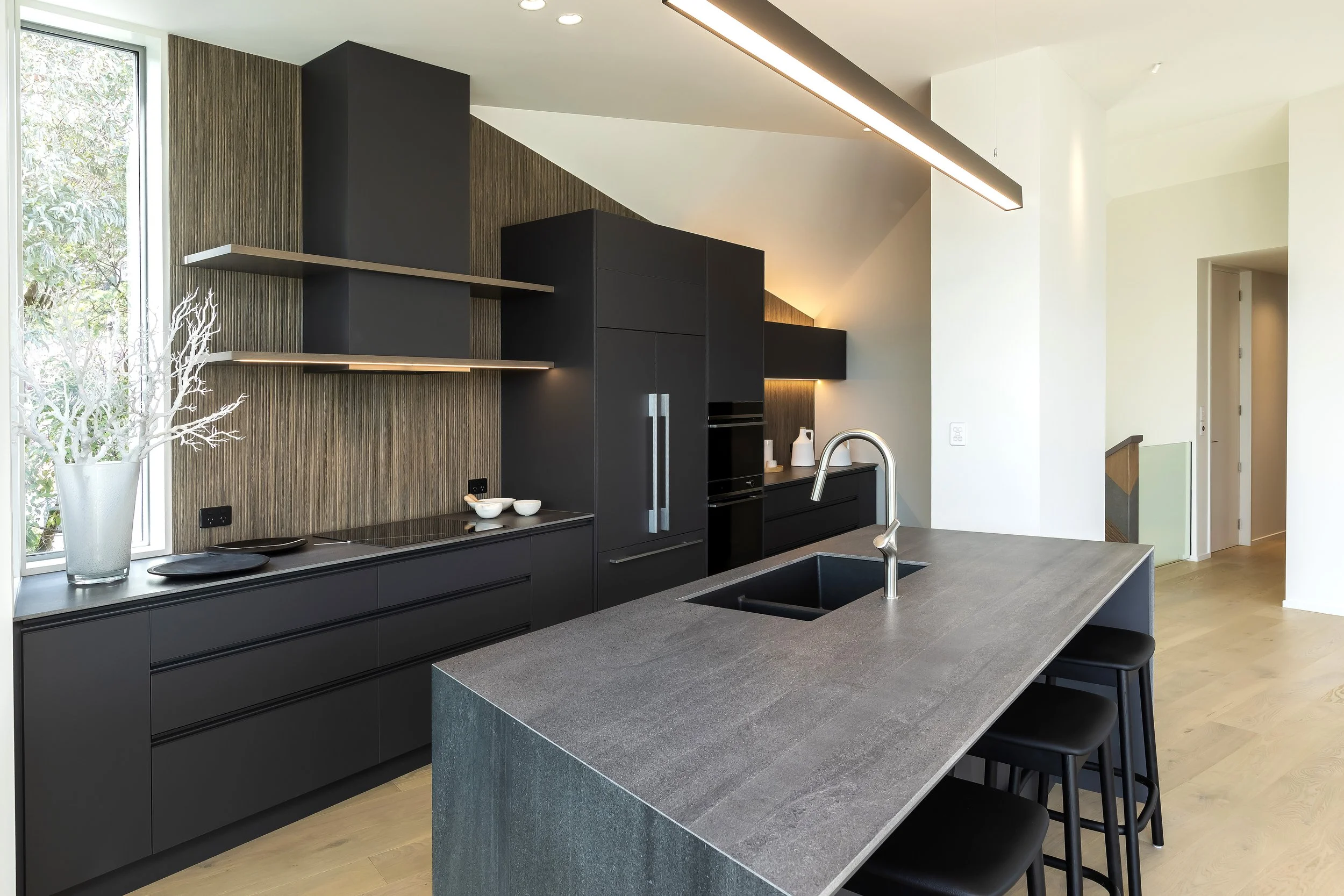 Modern kitchen with black cabinetry, a large kitchen island with a gray stone countertop, built-in appliances, a sink with a silver faucet, and a large window with a view of greenery. Kitchen Design by Bridget Coles Interior Design