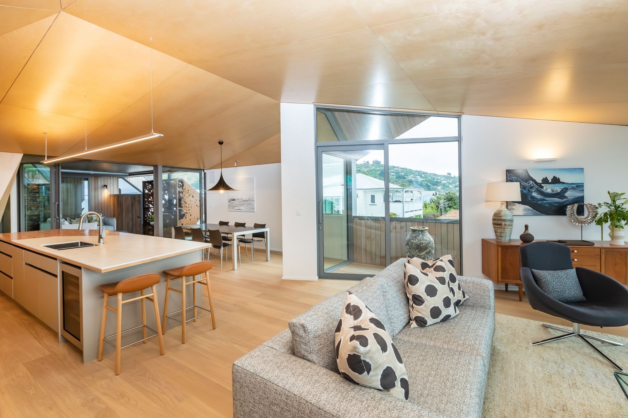 Modern open-concept living room and kitchen with wooden ceiling, large windows, and stylish furniture, including a sofa with polka dot pillows, a dining table, and a kitchen island with bar stools.