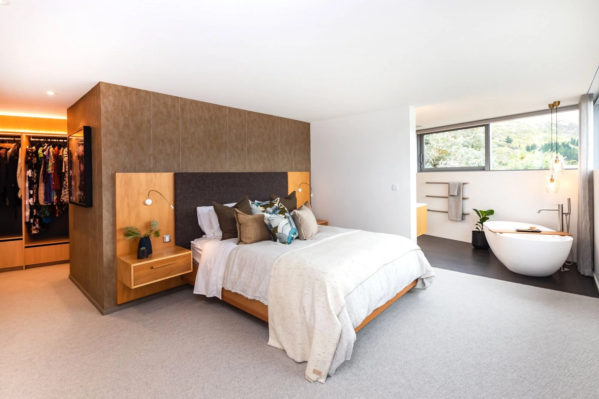Modern bedroom with a bed, pillows, bedside table, plants, and a bathtub near a large window with a view of greenery.