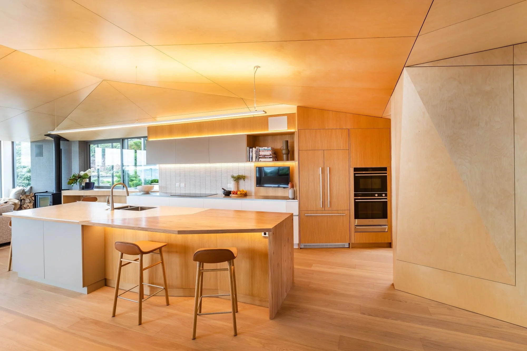 Modern kitchen with wooden cabinets, a white island with a wooden top, two bar stools, and stainless steel double ovens. Large windows and a ceiling with geometric wooden panels.