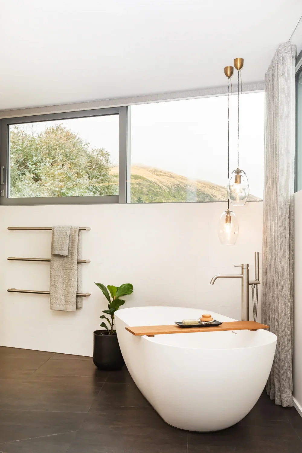 Modern bathroom with a free-standing oval bathtub, wooden tray with soap, towel rack with a towel, green plant in a black pot, large window showing trees and hills, pendant lights, and gray sheer curtains.