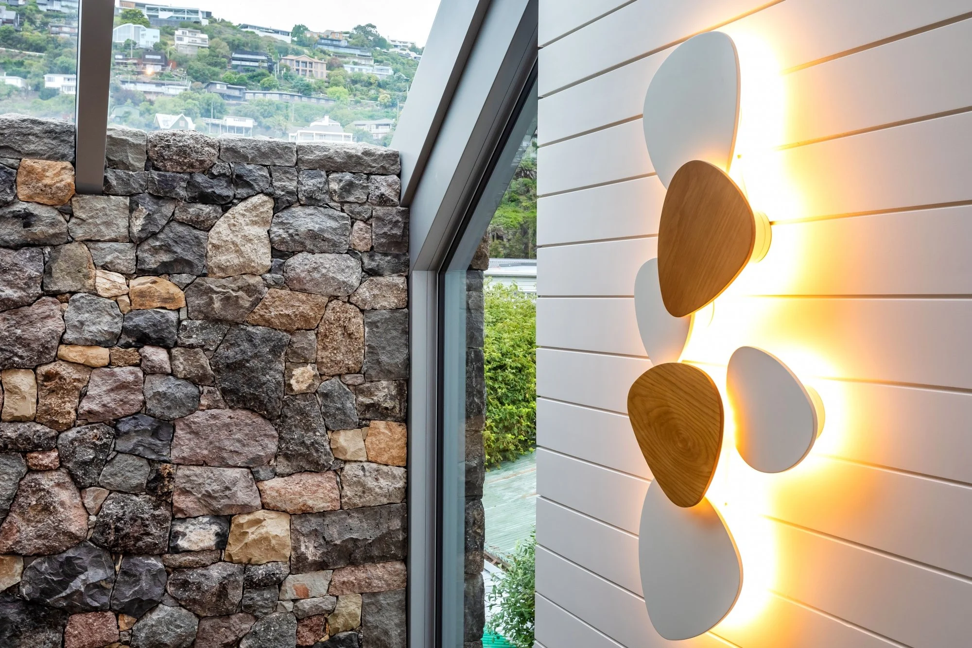 Interior of a building with a stone wall on the left, a large window showing a hillside with houses, and a white wood-paneled wall on the right with a wall-mounted light fixture that has multiple oval-shaped wooden and white elements emitting a warm 