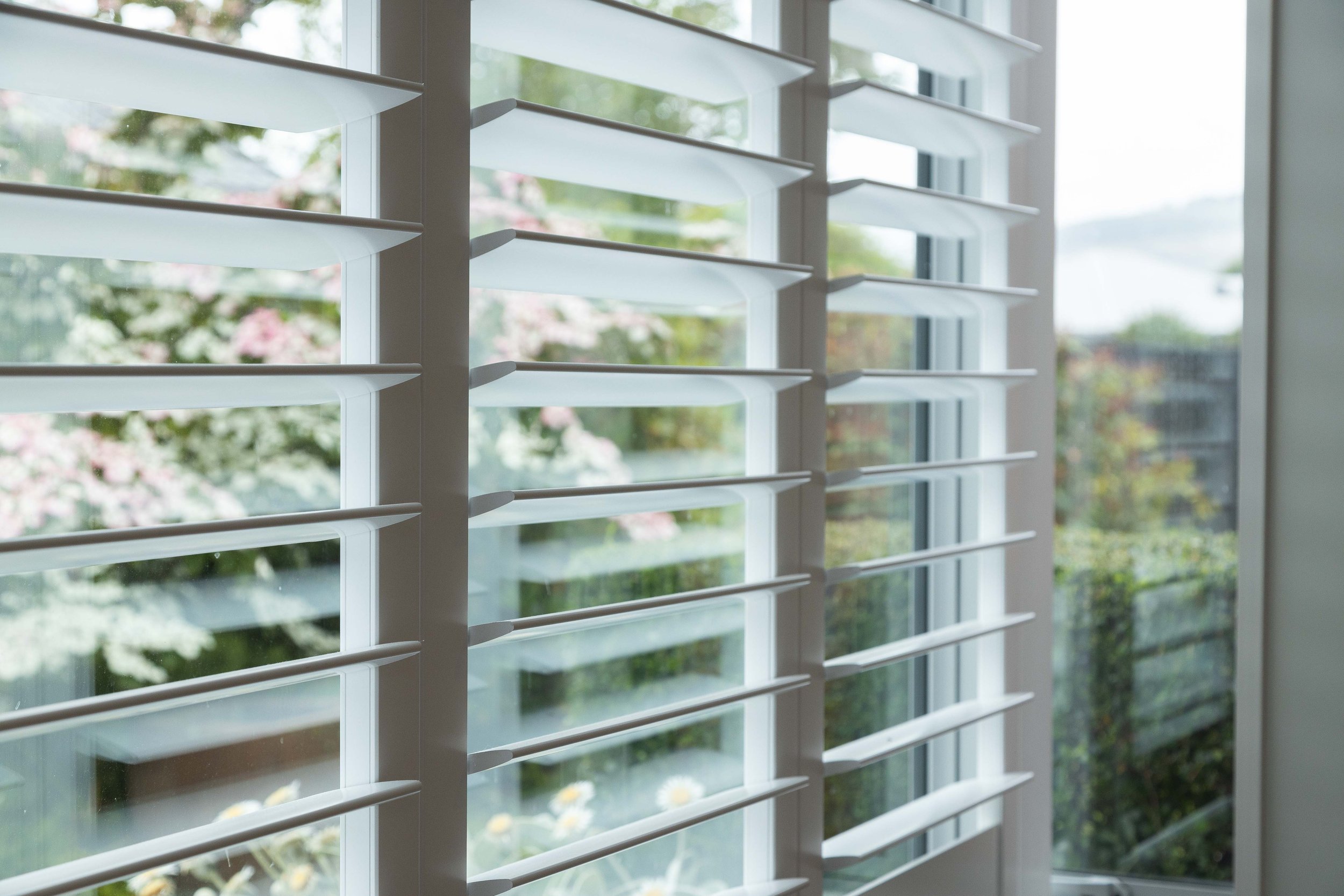 Close-up view of white window Norman shutters with a view of a garden outside through the window.