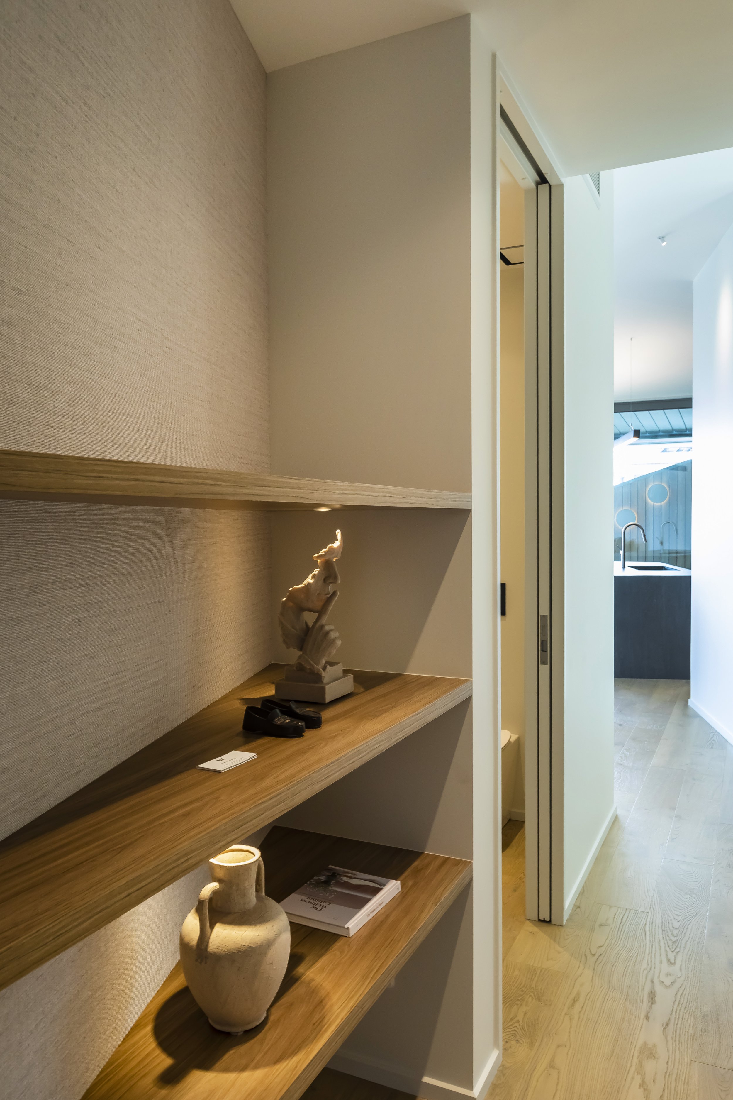 A wall with wooden shelves holding various decorative items, including a ceramic jug, a sculpture, and books, next to a hallway with wooden flooring and a sink visible in the background.