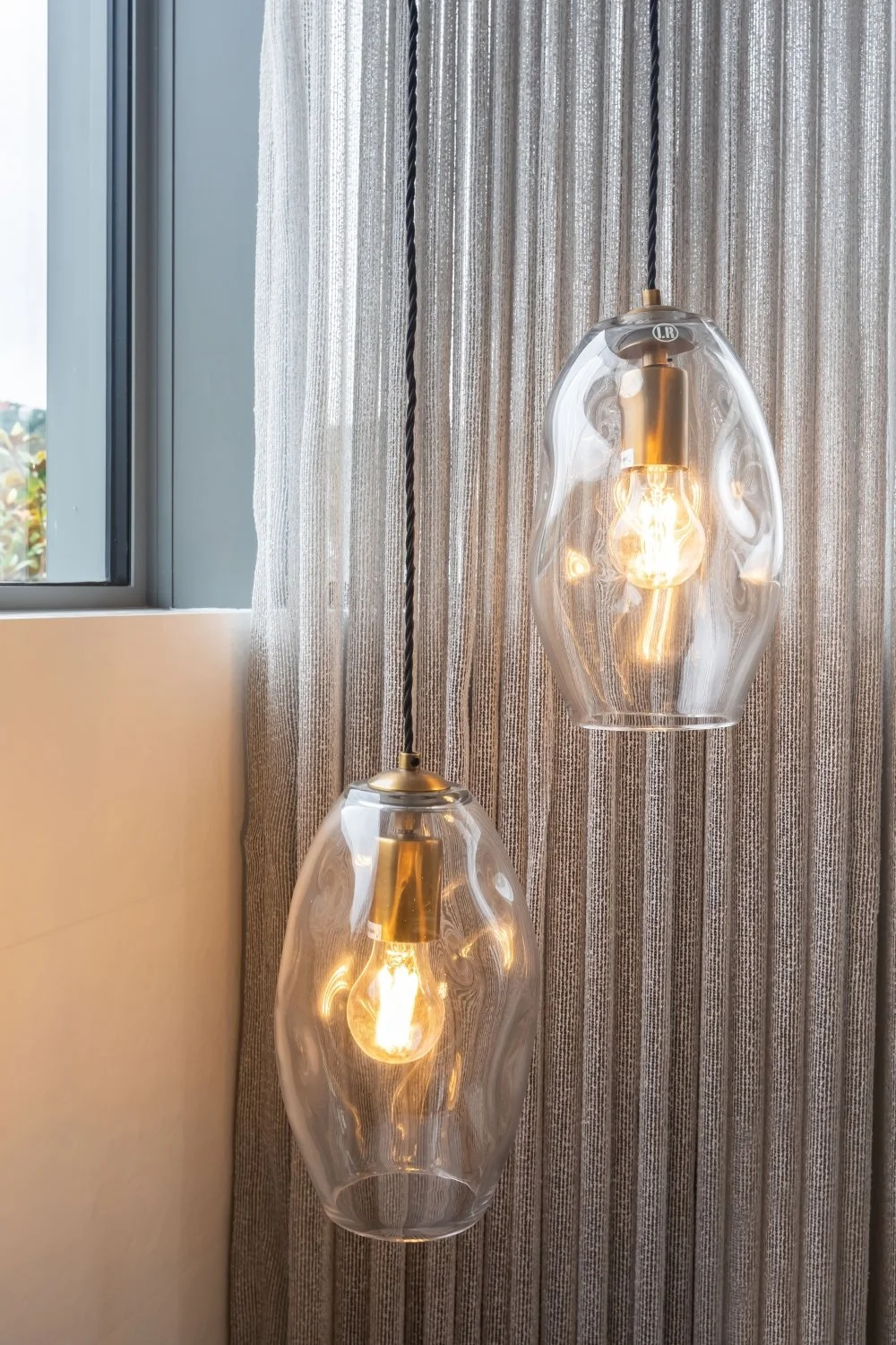 Two modern pendant lights with clear, irregularly shaped glass shades hanging indoors near a window with gray curtains.