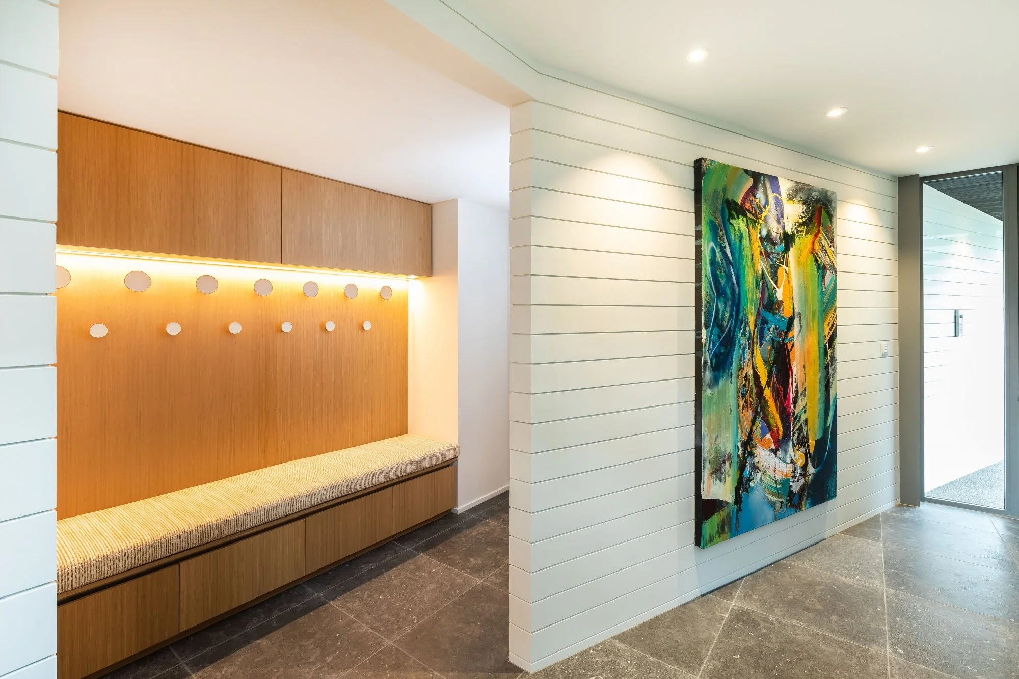 Interior view of a modern building lobby featuring a wooden bench with striped cushion, an abstract colorful painting on a white paneled wall.Joinery design by Bridget Coles Interior Design Christchurch BCSTUDIO