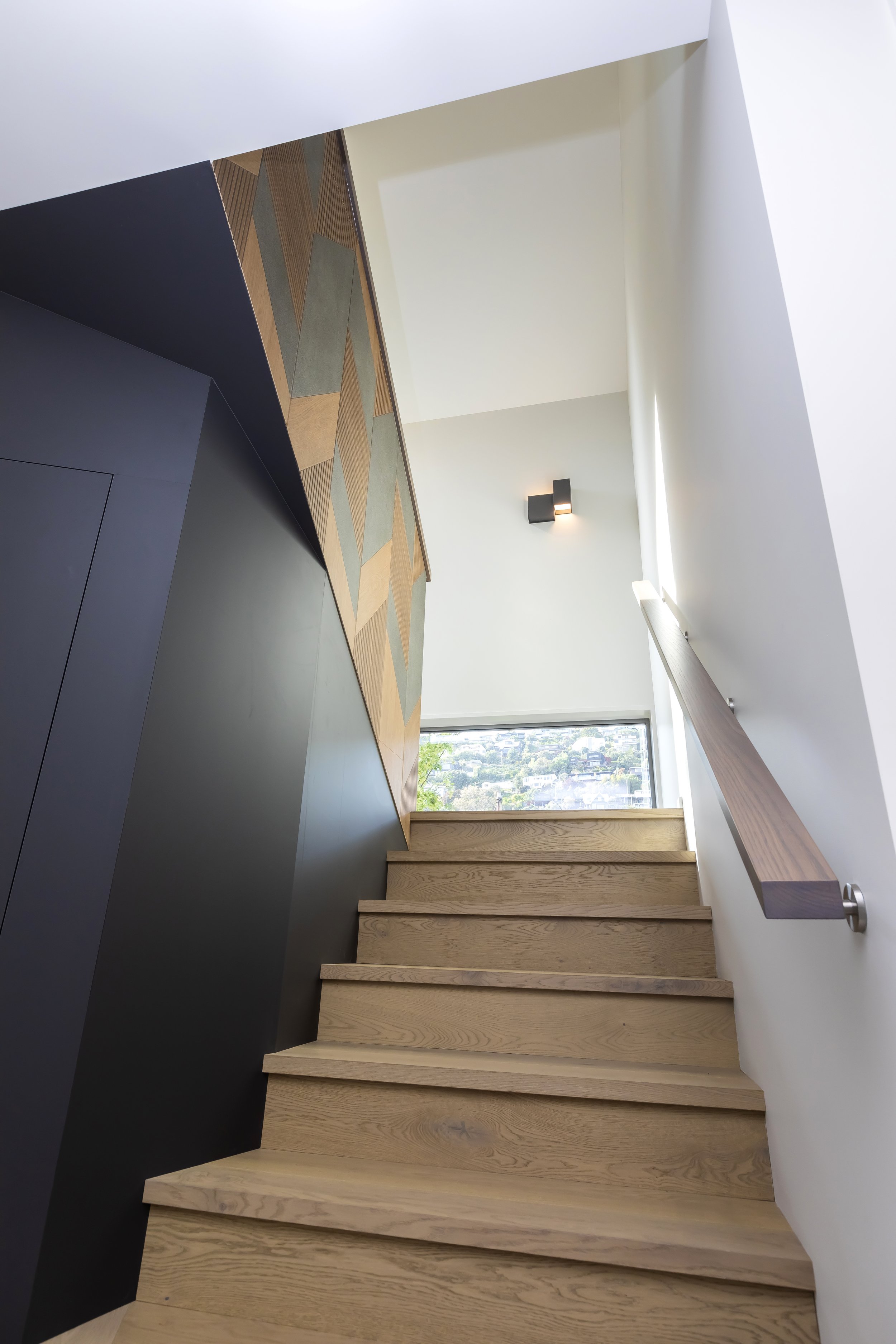 Interior shot of a modern staircase with wooden steps, a wooden handrail, and a large window allowing natural light to illuminate the space. Stair Design by Bridget Coles Interior Design