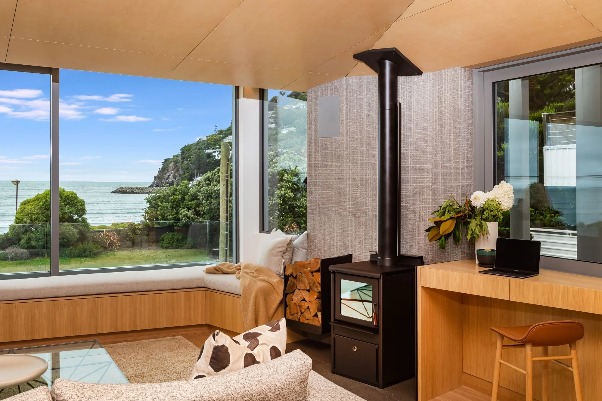 Living room with large windows showing a view of the ocean and greenery, a wood-burning stove, flowers on a table, and modern furniture.