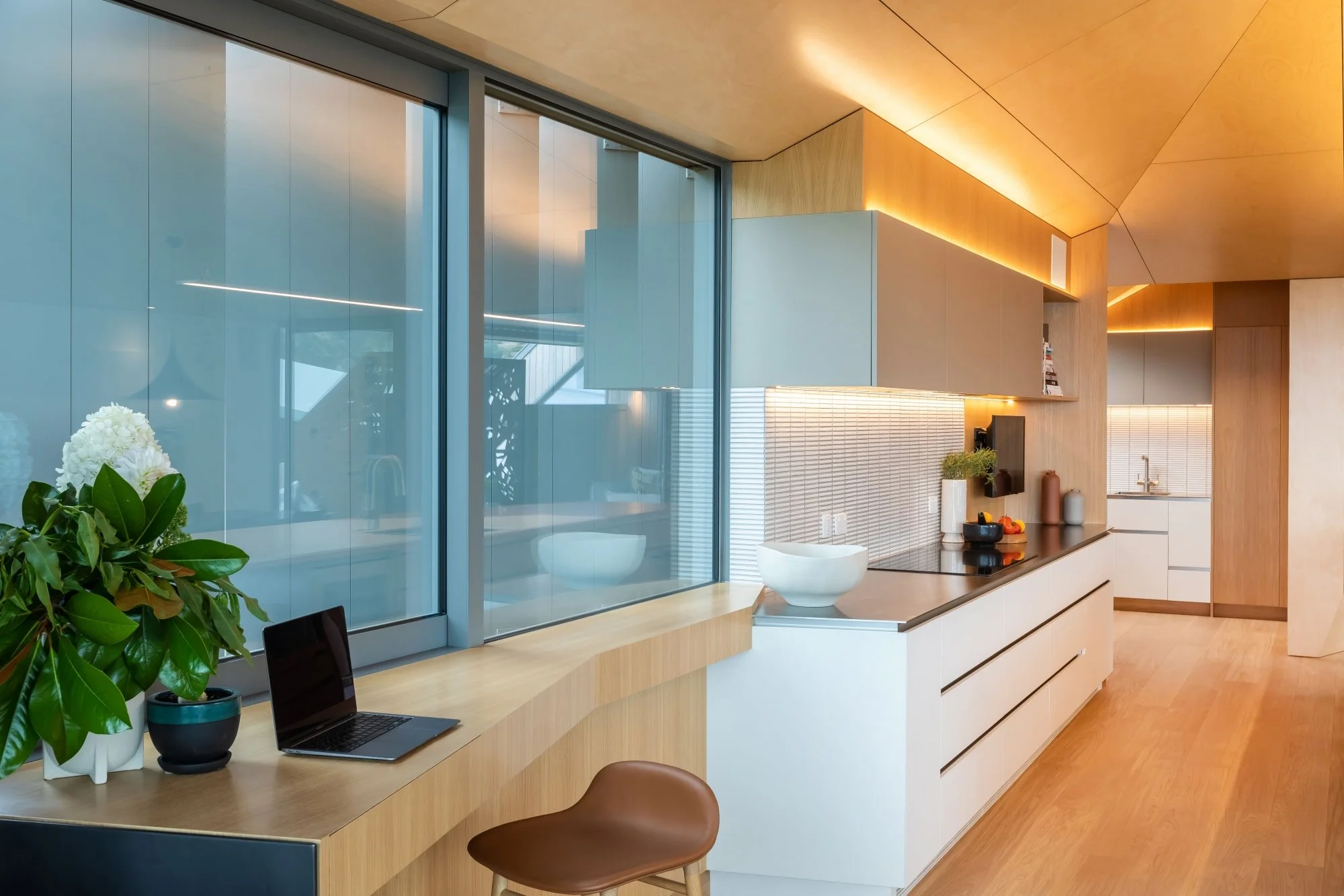 Modern kitchen with wooden cabinets, a countertop with decorative vases, and a large window with frosted glass panels. There is a laptop, a potted plant, and a chair at a wooden table.