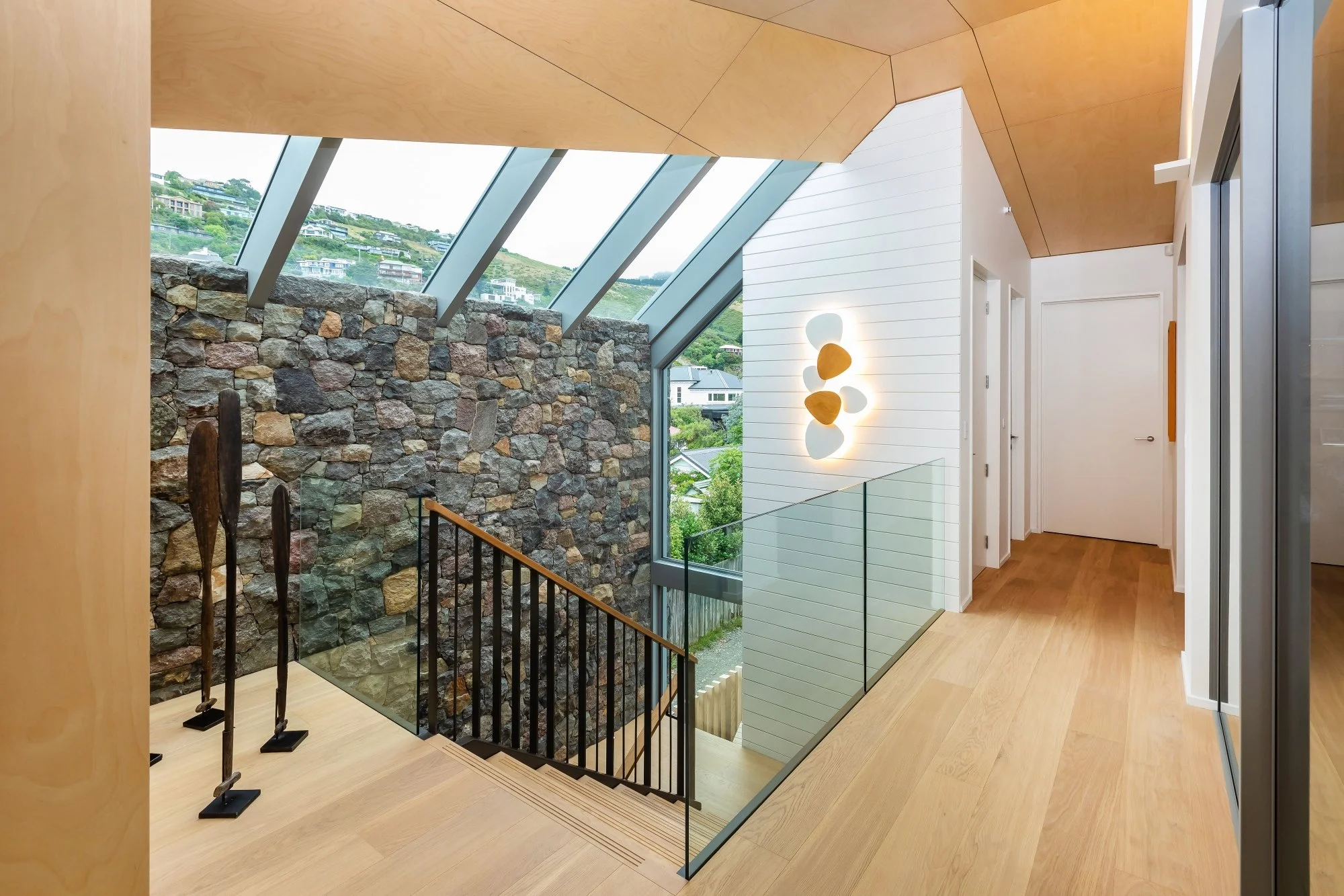 Interior view of a modern house with large sloped glass windows, a stone accent wall, wooden ceiling and flooring, and a hallway with white walls and a light fixture.Interior design Bridget Coles Interior Design Christchurch BCSTUDIO