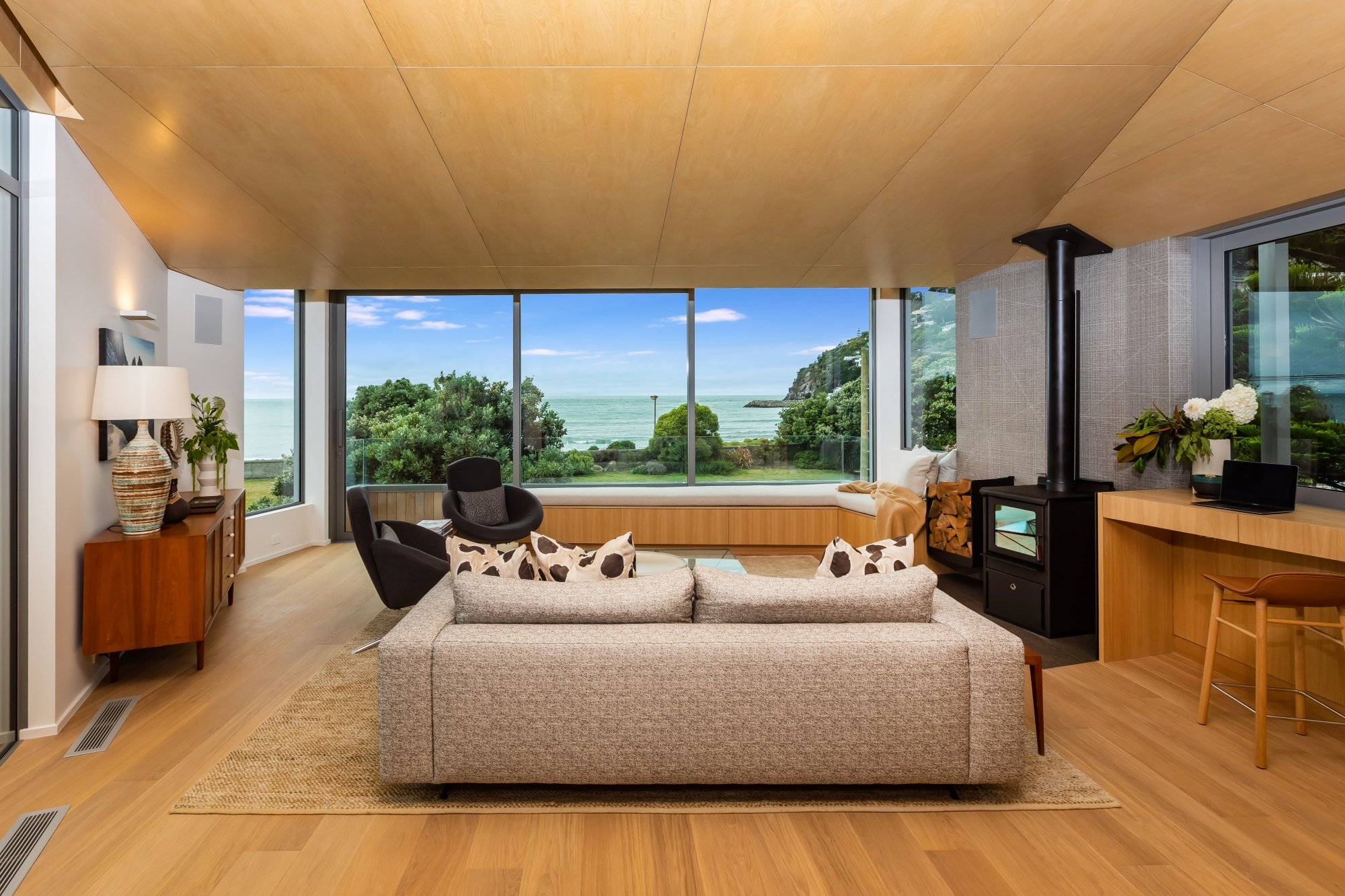 Coastal Living room with large windows showing a view of greenery and the ocean, with a beige sofa, black armchair, wooden side tables, and a wood stove.